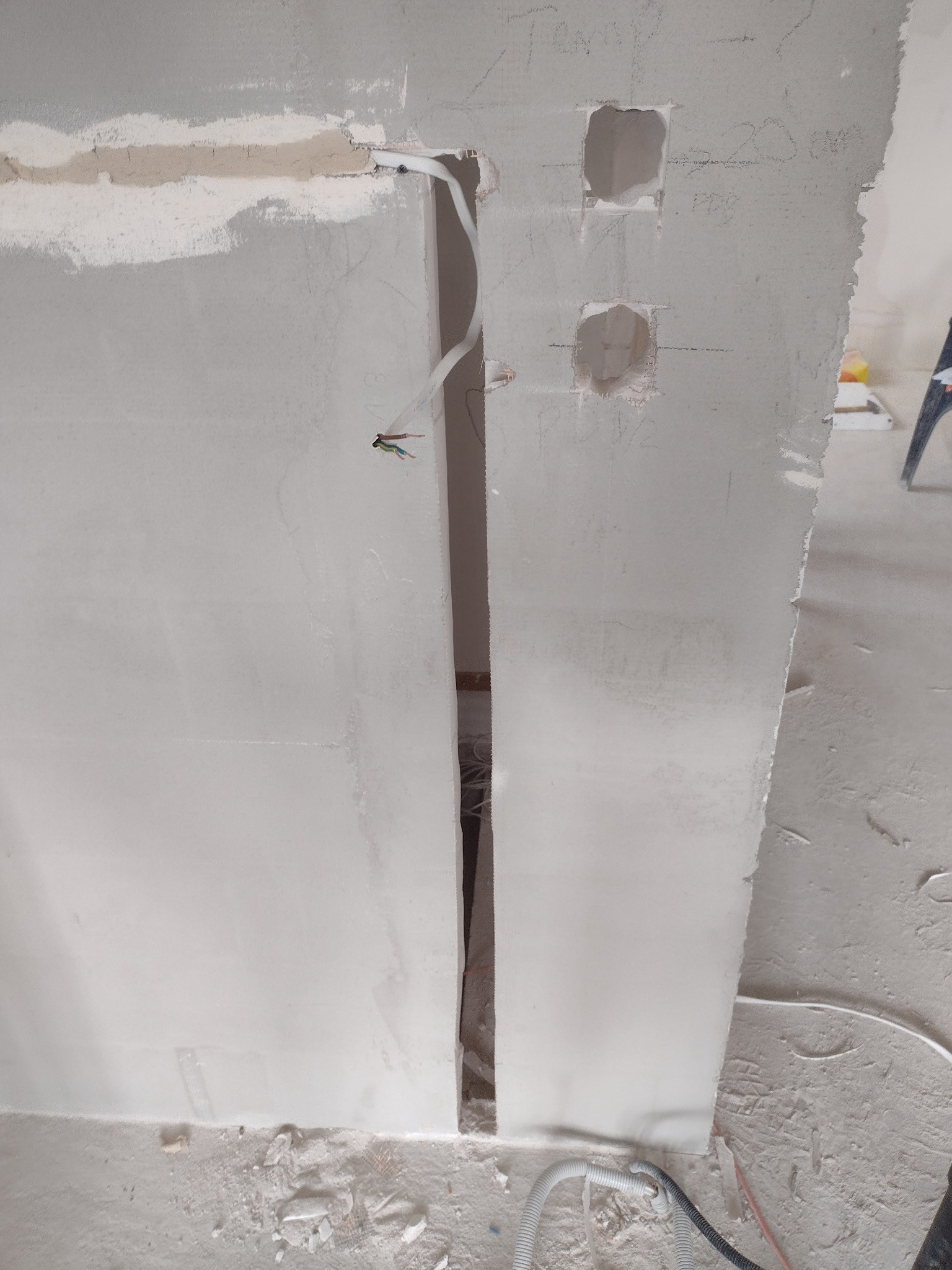 Ściana w trakcie prac wykończeniowych, z widocznymi kablami elektrycznymi wychodzącymi ze ściany, otwory na gniazdka i fragmenty zagruntowanej powierzchni.