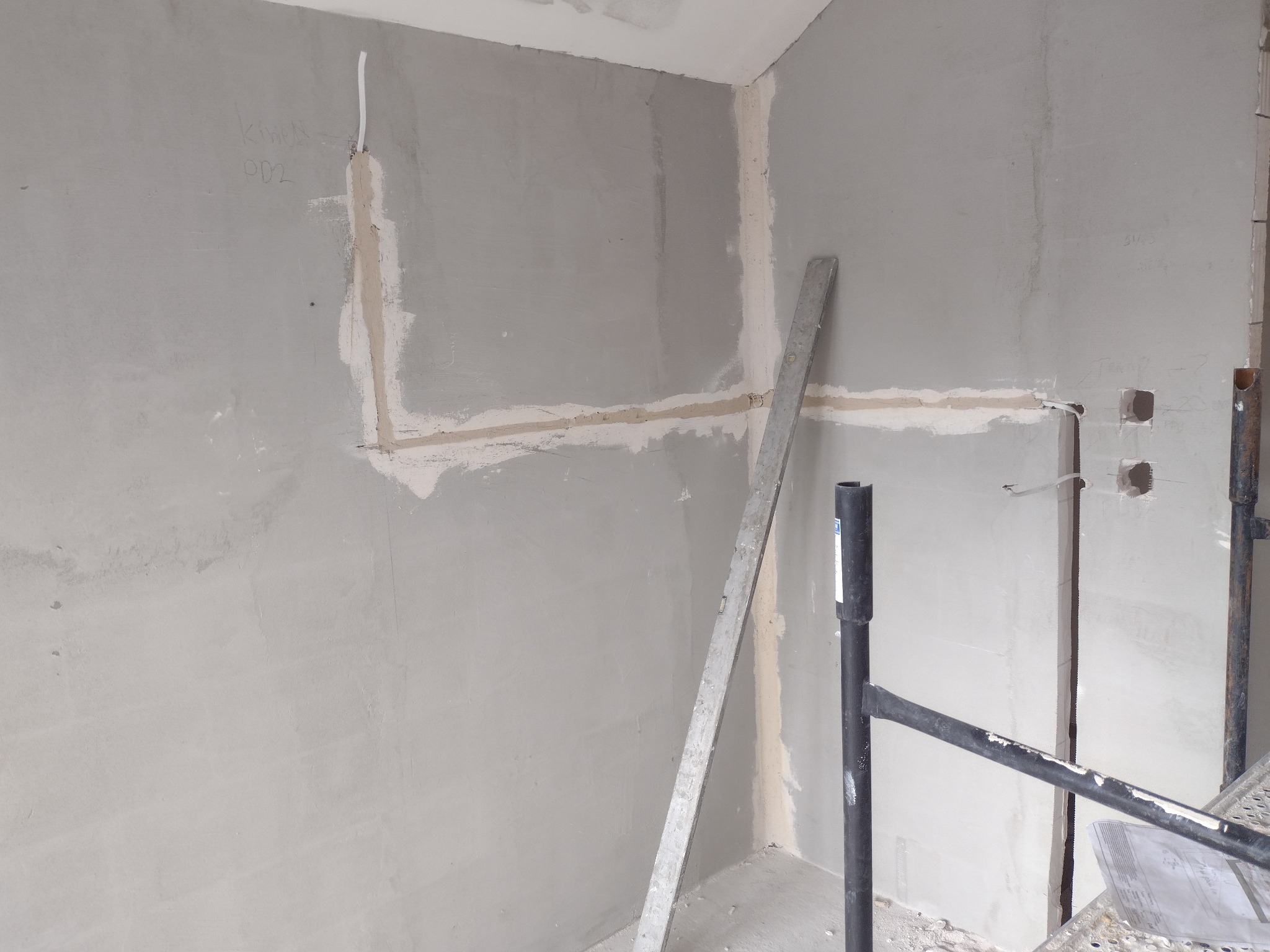 Ściana w trakcie prac przygotowawczych do wykończenia, widoczne ślady po instalacjach elektrycznych, częściowo wypełnione bruzdy, pionowa poziomica oparta o ścianę, fragment rusztowania.