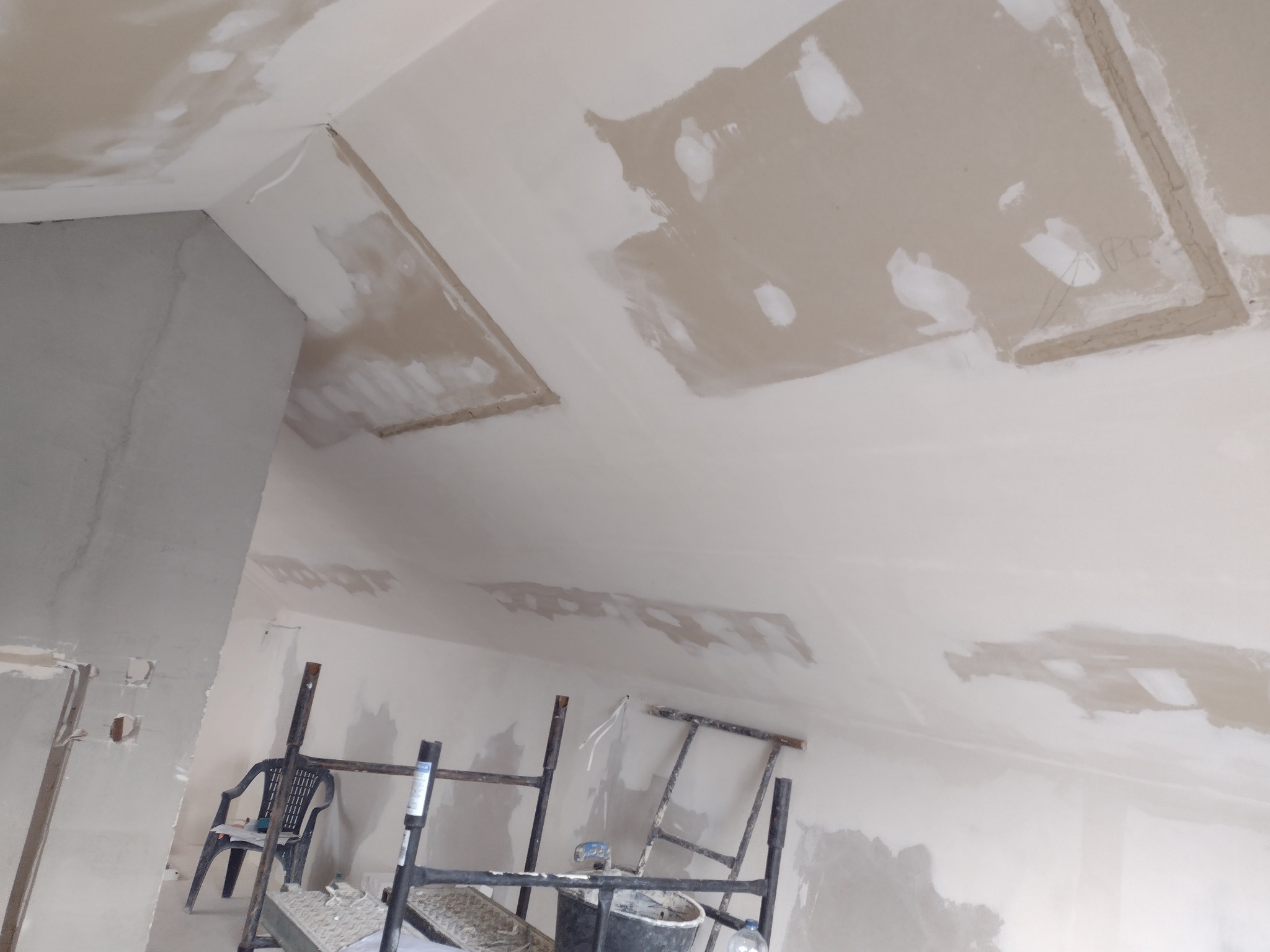 Sufit w trakcie prac wykończeniowych z widocznymi łatami i nierównościami, ustawione rusztowanie i krzesło w pomieszczeniu, przygotowania do dalszych prac.
