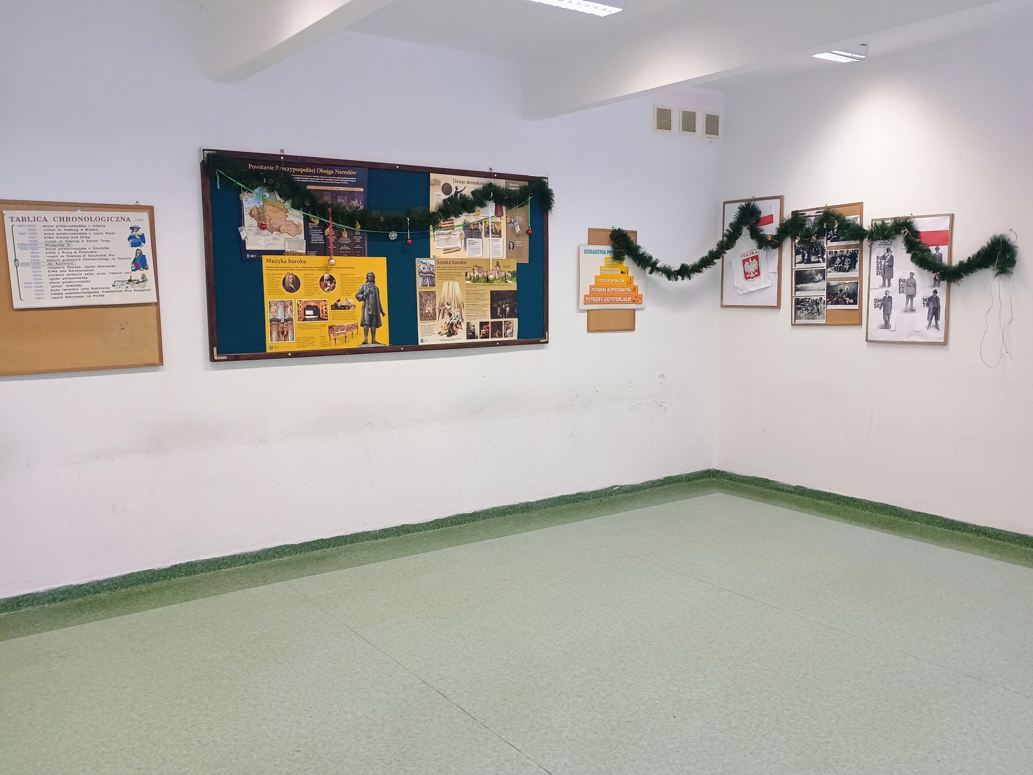 Korytarz szkolny z tablicami tematycznymi ozdobionymi świątecznymi girlandami, widok z niskiej perspektywy ukazujący podłogę i dolną część ścian.