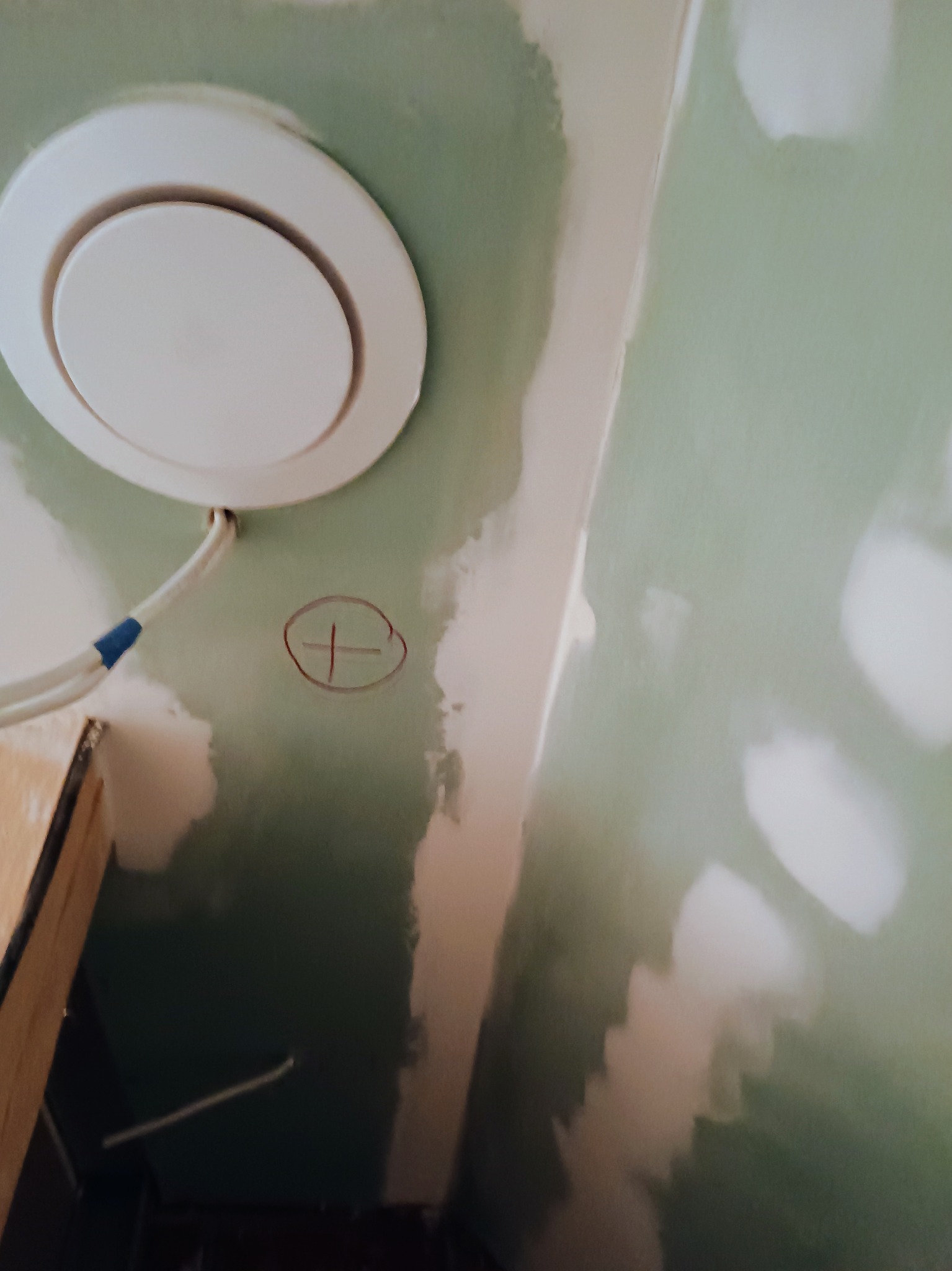 Niedokończona ściana z płyt kartonowo-gipsowych w trakcie szpachlowania, widoczny okrągły nawiewnik wentylacyjny z podłączonymi kablami i oznaczenie krzyżykiem na ścianie.