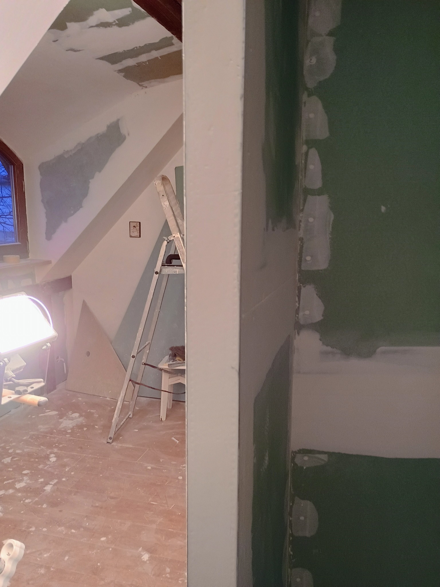 Pokój w trakcie remontu: widoczna ściana z zielonej płyty karton gips, łączenia szpachlowane, w tle drabina i lampa budowlana.