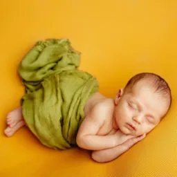 Śpiący noworodek owinięty w zieloną tkaninę na żółtym tle, dłonie pod policzkiem, widoczne małe stopy.