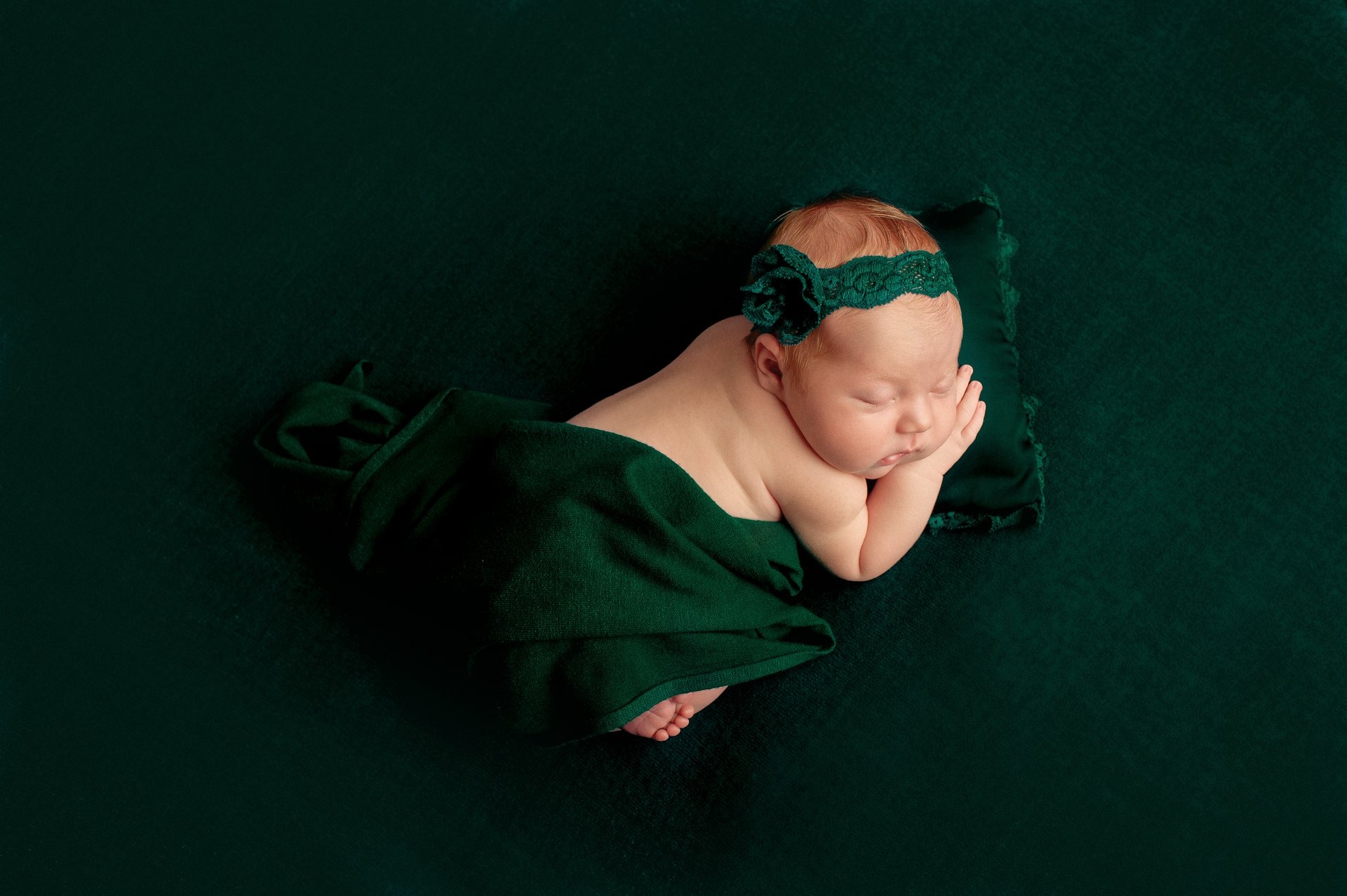 Śpiąca noworodek owinięta w zielony materiał, z zieloną koronkową opaską na głowie, leżąca na zielonej poduszce i tle.