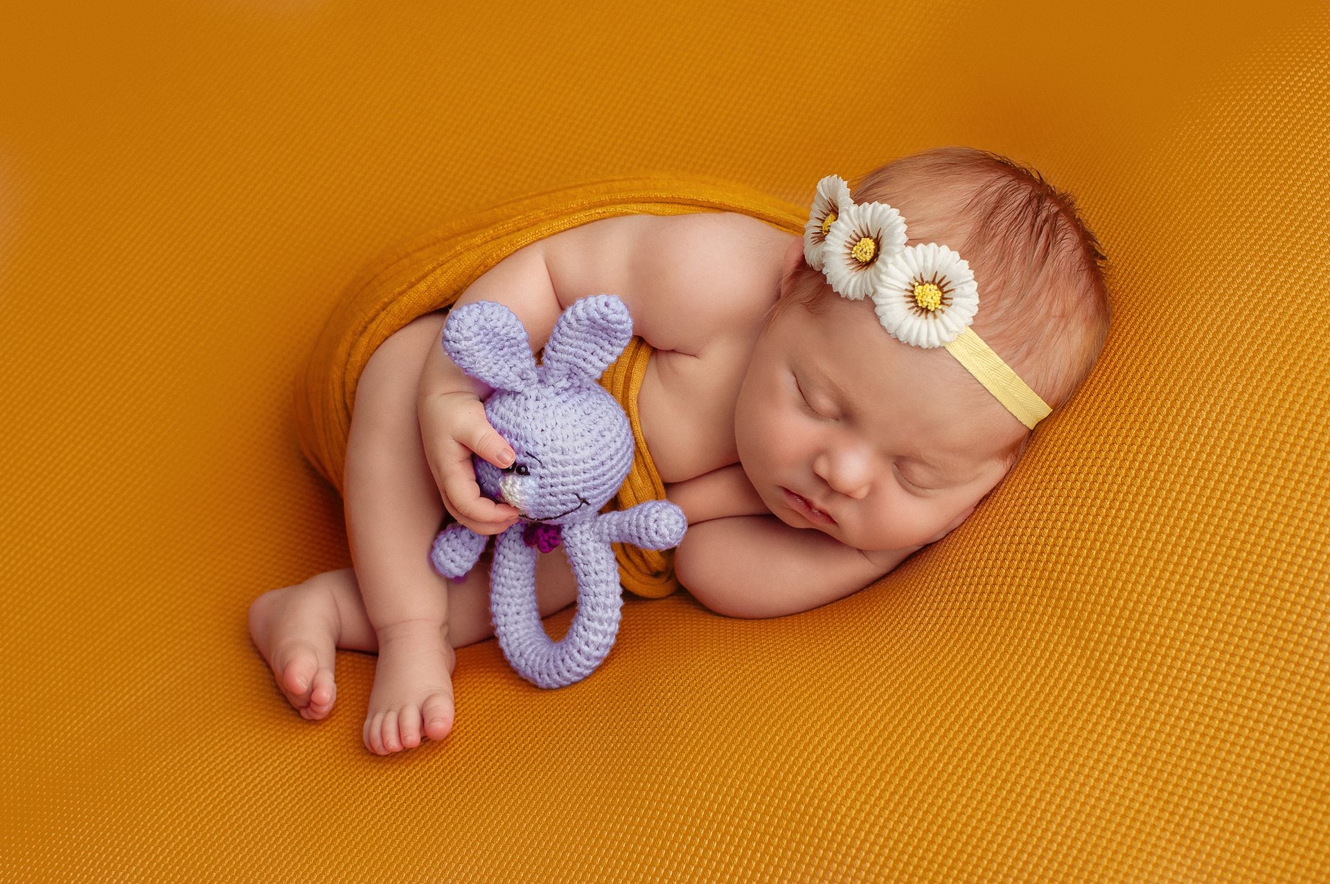 Noworodek śpiący na pomarańczowym tle, otulony żółtym materiałem, trzymając w rączce fioletową, dzierganą zabawkę-gryzak w kształcie królika, z opaską z białych kwiatów na głowie.