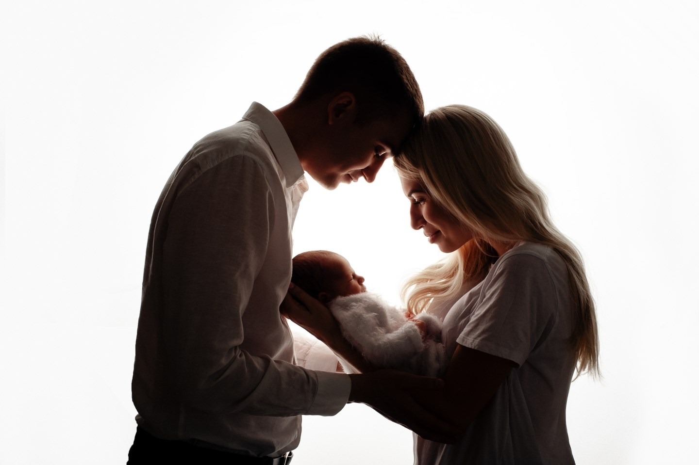 Silhouettes of a young couple holding their newborn baby, heads touching, soft white background.