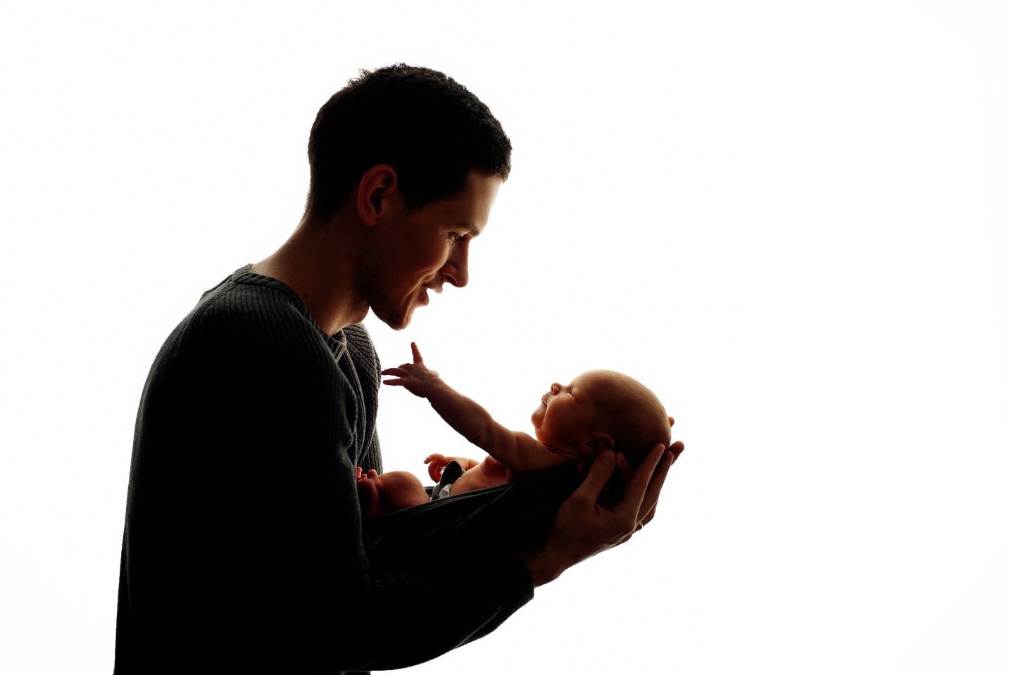 Silhouetted father holding newborn baby, looking at infant's outstretched hand against a bright white background.