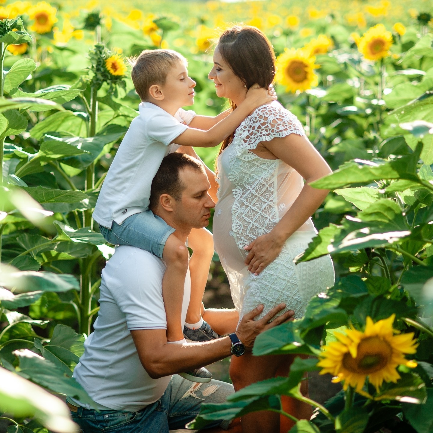 Ciężarna kobieta w białej sukience, tulona przez syna siedzącego na ramionach ojca, na tle pola słoneczników w słoneczny dzień.