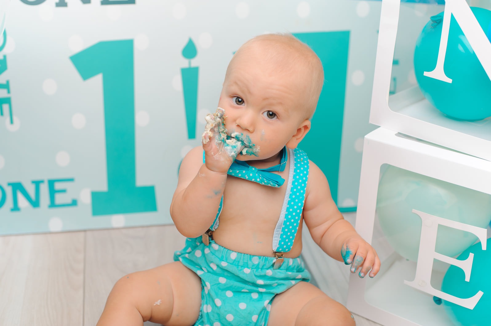 Uroczy roczny chłopiec w turkusowej muszce i spodenkach w kropki, siedzi na podłodze z ciastem na twarzy i palcach, świętując pierwsze urodziny na tle dekoracji z napisem 'ONE'.