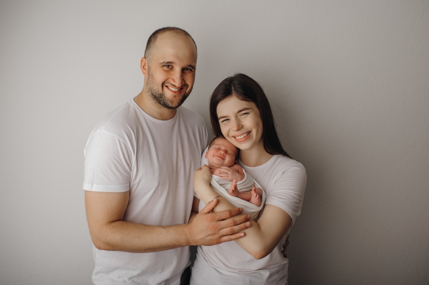 Szczęśliwa młoda rodzina: uśmiechnięty ojciec, matka trzymająca zawinięte w biały materiał nowo narodzone dziecko z zamkniętymi oczami, wszyscy ubrani w białe T-shirty na jasnoszarym tle.