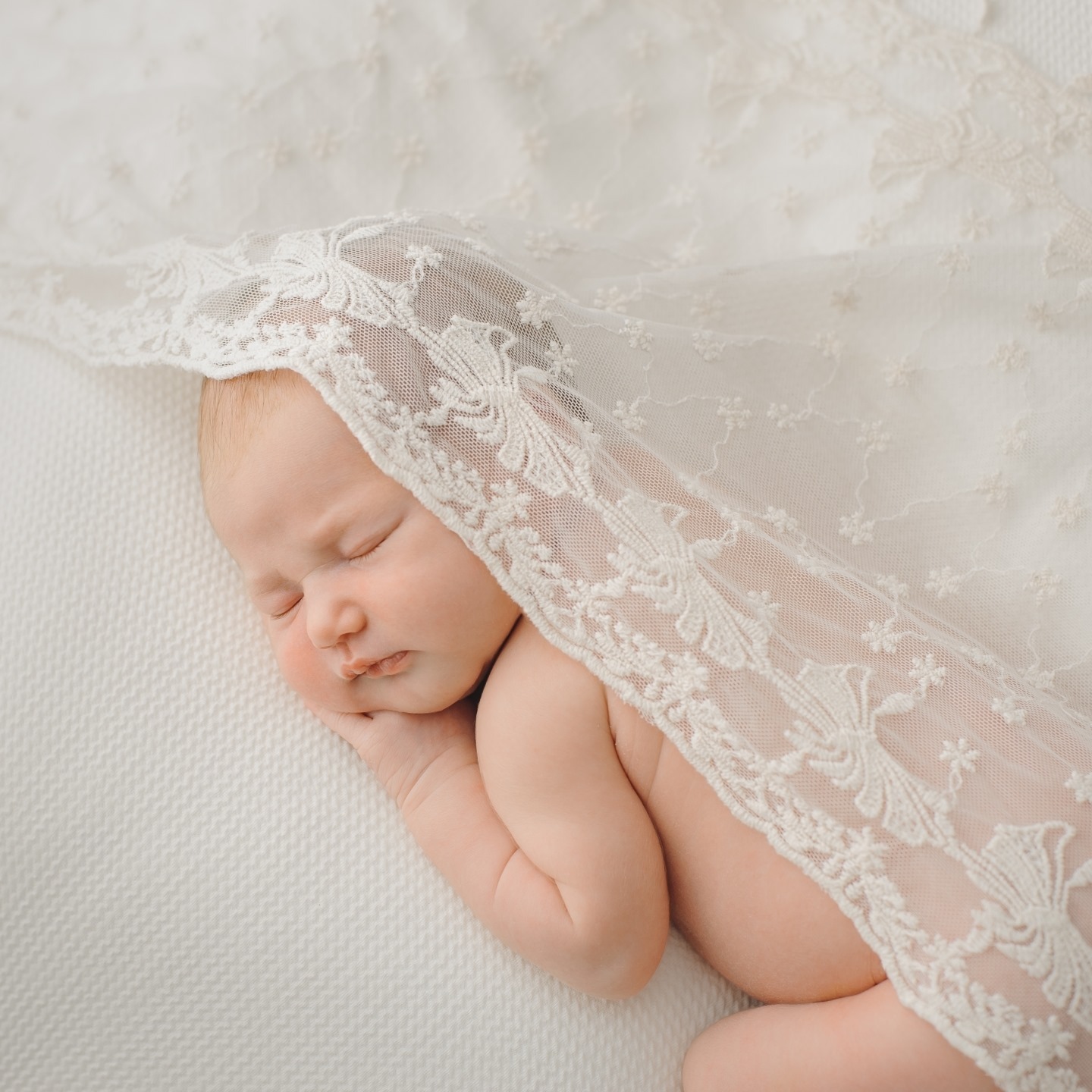 Śpiący noworodek owinięty w delikatną, kremową koronkę na białym teksturowanym tle, z subtelnym oświetleniem podkreślającym jego delikatną skórę.