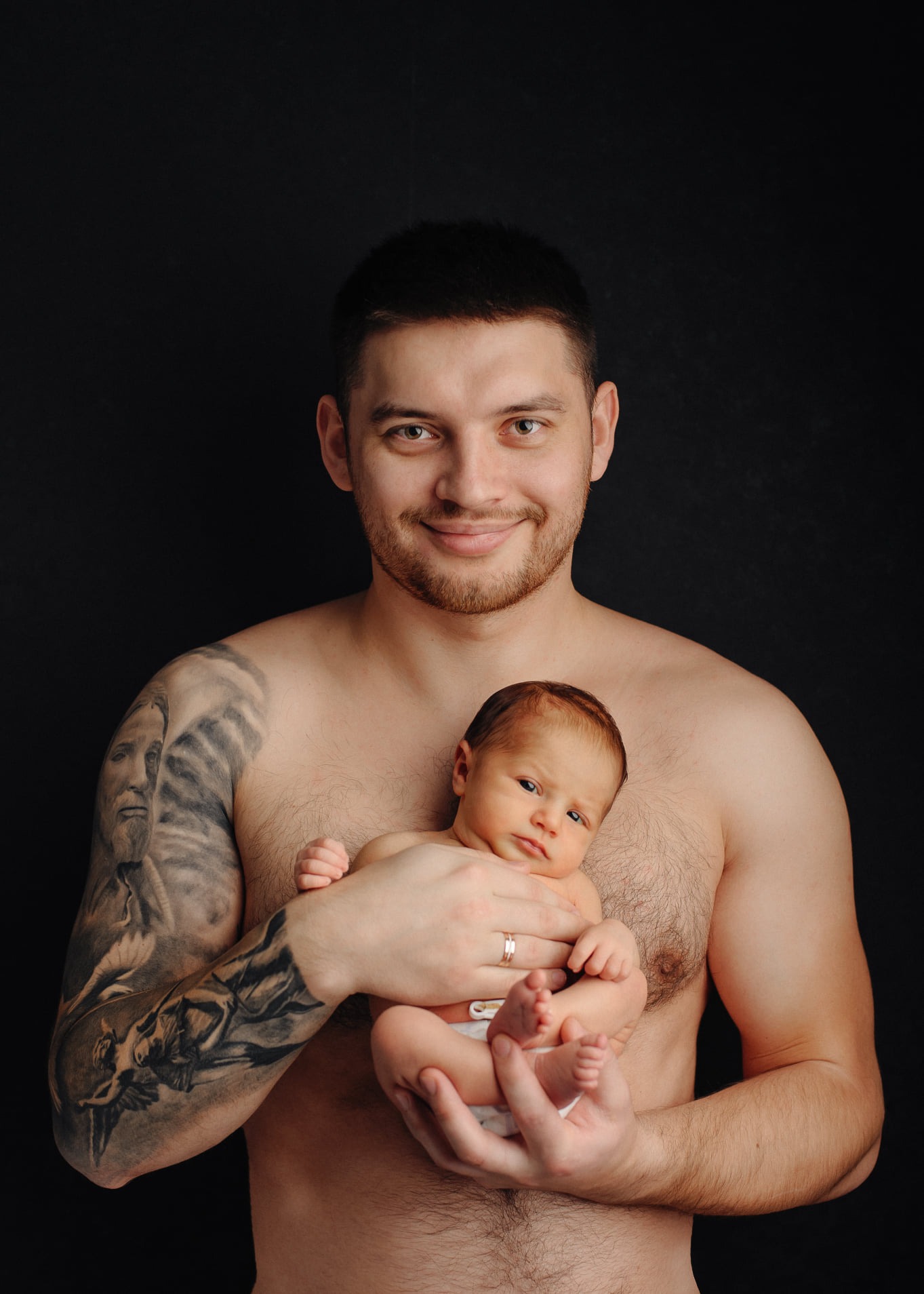 Uśmiechnięty, brodaty mężczyzna z tatuażem na ramieniu trzyma noworodka na tle czarnego tła.
