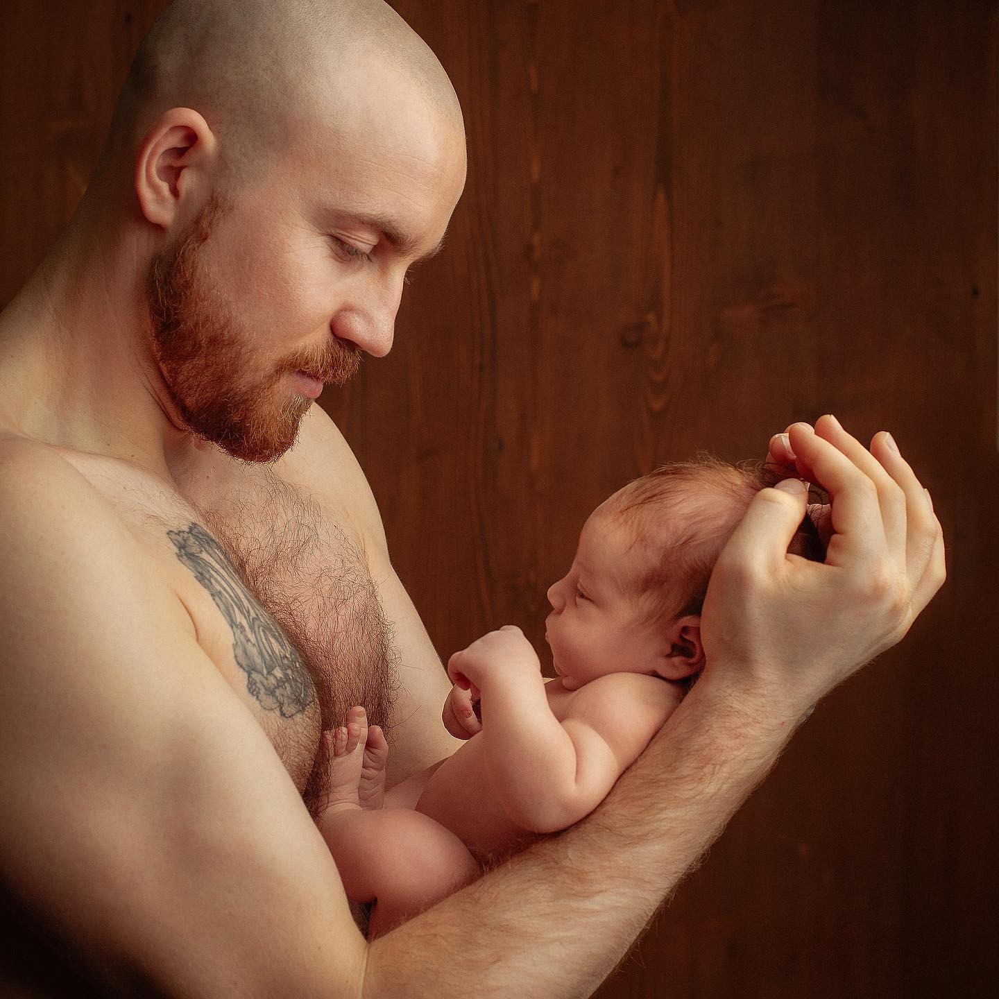 Ojciec z tatuażem na ramieniu trzyma delikatnie noworodka na tle drewnianej ściany, oboje patrzą na siebie.