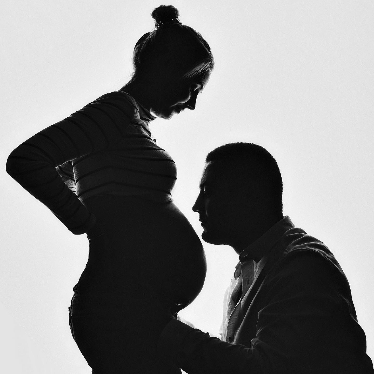 Silhouetted pregnant woman in striped top with partner touching her belly, monochrome studio shot.