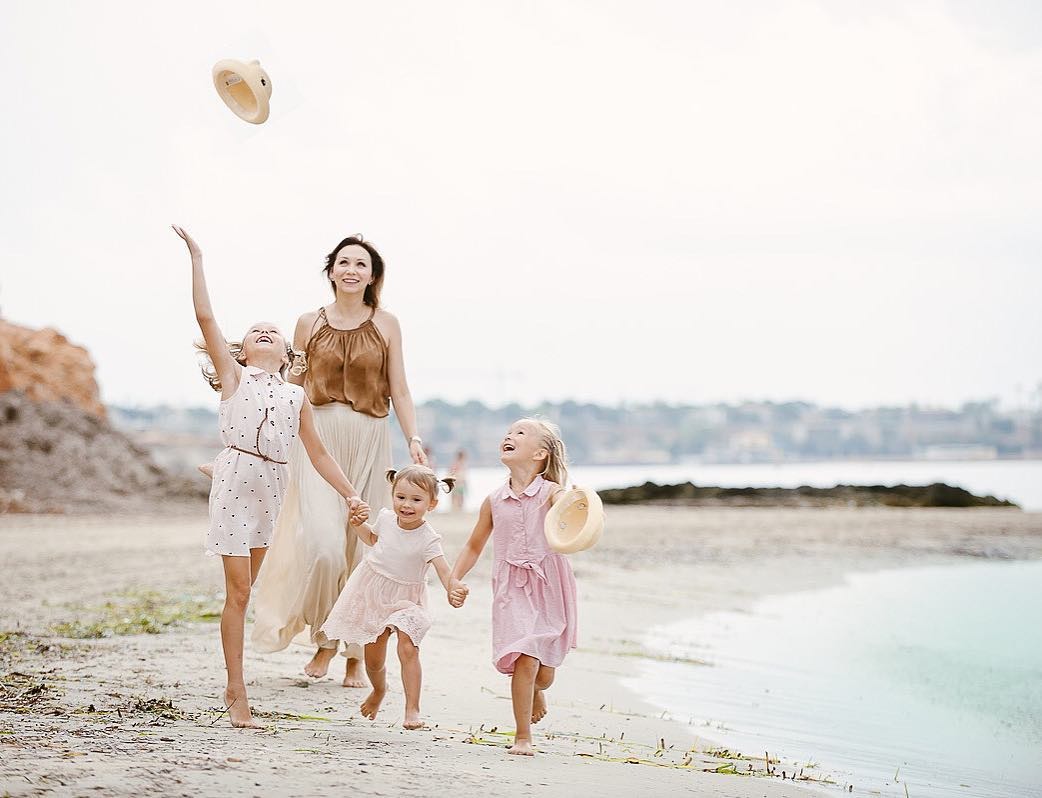 Kobieta z trzema córkami spaceruje po plaży, jedna z dziewczynek podrzuca słomkowy kapelusz, pozostałe dzieci trzymają się za ręce i biegną wzdłuż brzegu morza.