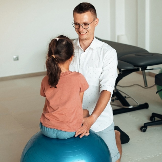 Uśmiechnięty mężczyzna w okularach pomaga dziewczynce siedzieć na dużej, niebieskiej piłce do ćwiczeń w gabinecie, w tle widoczny stół do rehabilitacji.