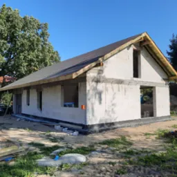 Surowy stan domu z białych bloczków, widoczne fundamenty z izolacją, konstrukcja dachu z membraną, w tle drzewa i samochody.