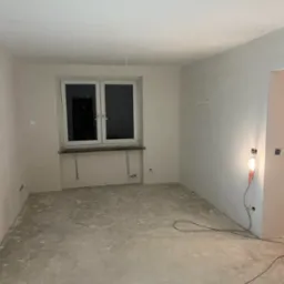 Pokój w trakcie remontu, ściany pokryte gładzią, widoczne przewody elektryczne, surowy beton na podłodze, prowizoryczne oświetlenie.