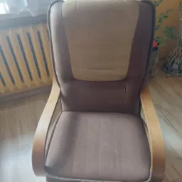 Brązowy fotel z jasnymi wstawkami, widoczne drewniane podłokietniki i fragment kaloryfera w tle.