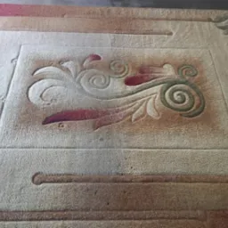 Beżowy dywan z bordowo-zielonym ornamentem roślinnym, widoczne ślady użytkowania i potencjalne zabrudzenia.