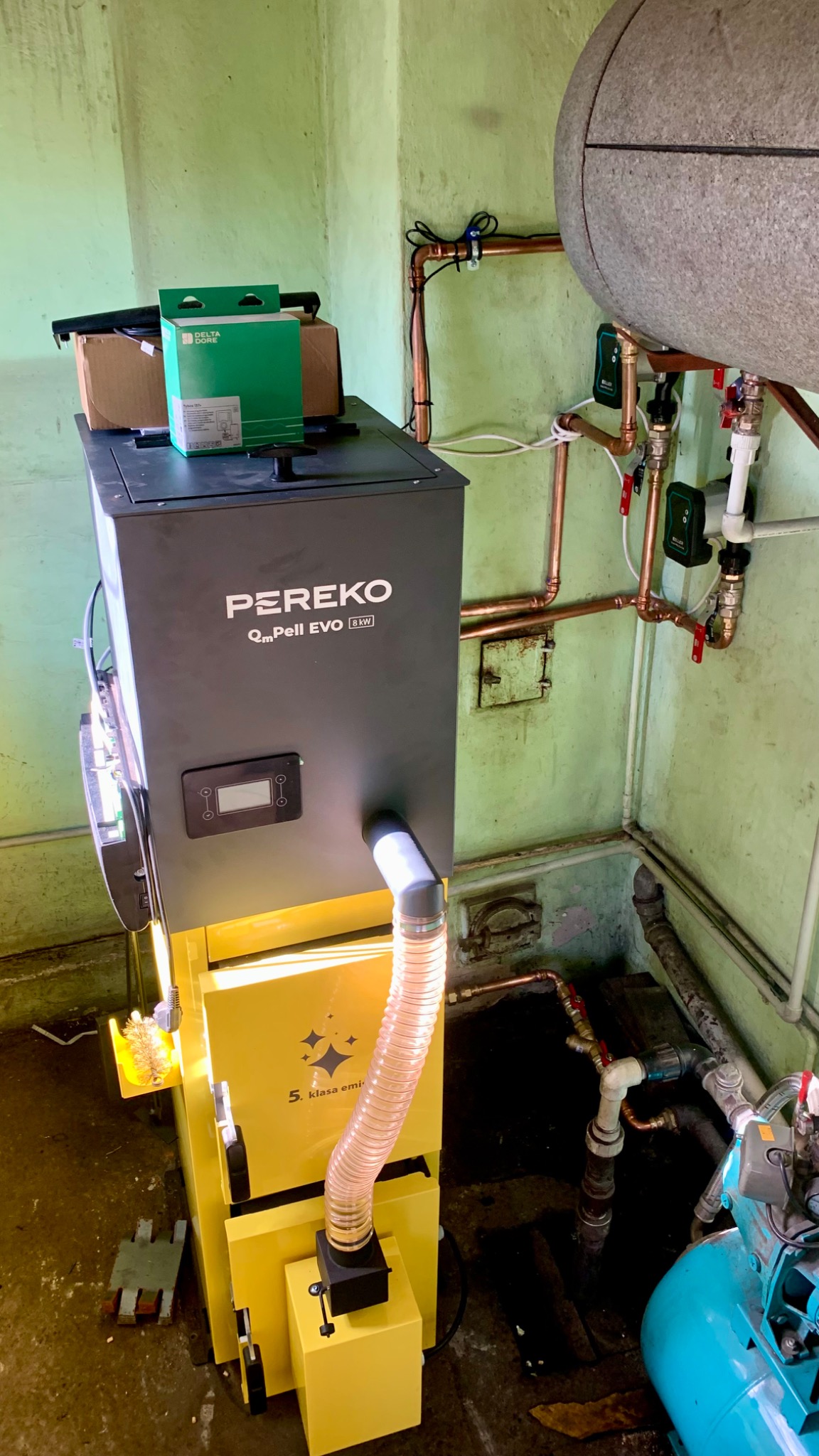 Żółto-czarny piec Pereko QmPell EVO 8kW z podajnikiem, widoczne rury miedziane instalacji CO i zbiornik buforowy w kotłowni o zielonych ścianach.