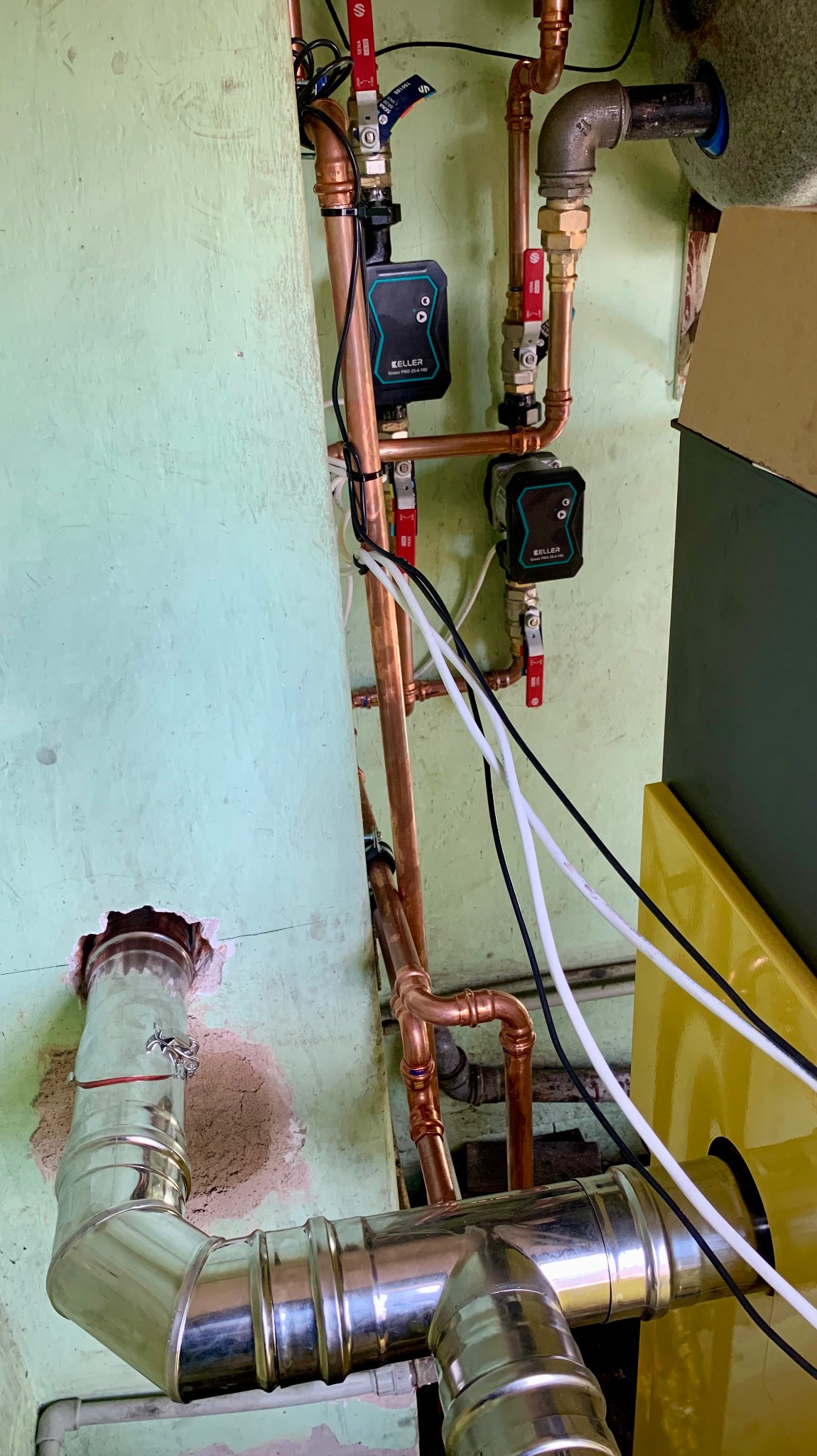 Fragment instalacji grzewczej z miedzianymi rurami, pompami obiegowymi Keller Smart PRO 25-4-180 oraz stalowym kominem spalinowym podłączonym do żółtego kotła, na tle ściany o zielonkawym odcieniu.
