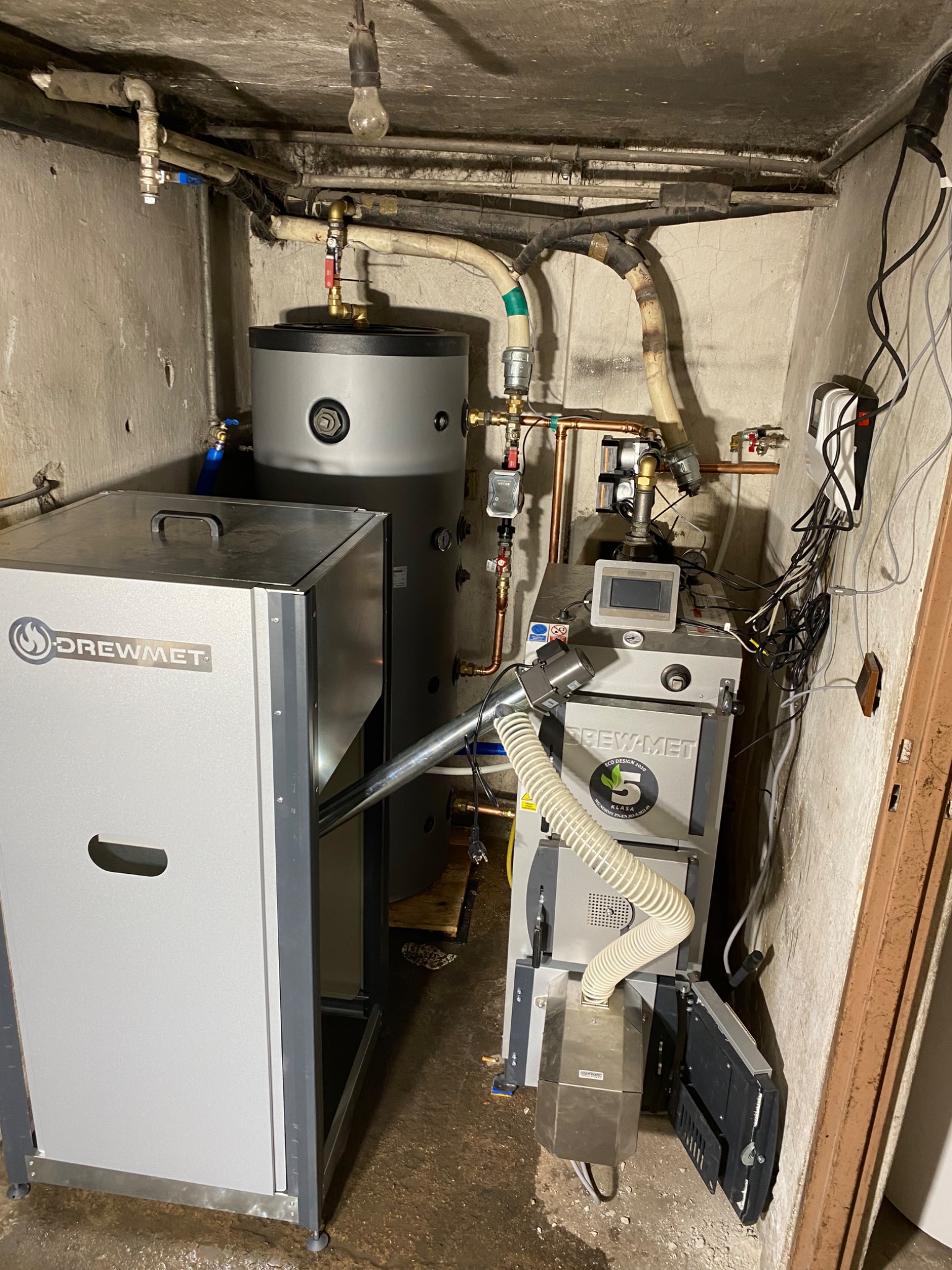 Zestaw kotła na paliwo stałe marki Drewmet z zasobnikiem ciepłej wody w kotłowni. Widoczne rury instalacji grzewczej i sterownik kotła. Narożne ujęcie.
