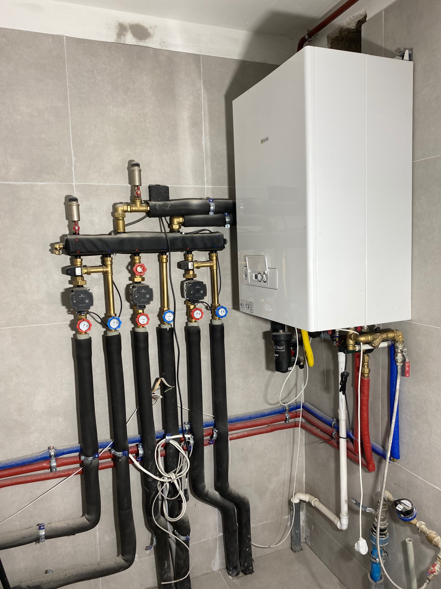 Biały piec gazowy Beretta z czarnymi rurami i zaworami na szarej, kafelkowanej ścianie. Widoczne rury miedziane i plastikowe, instalacja w kotłowni.