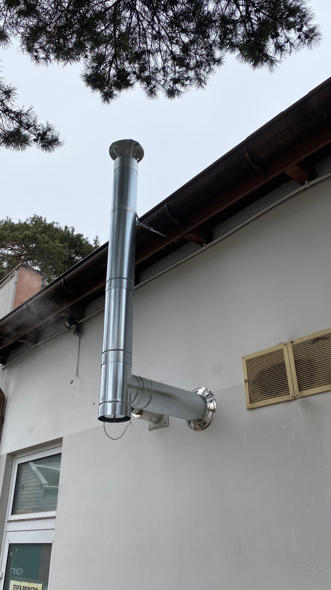 Zewnętrzny, stalowy komin spalinowy zamontowany przy ścianie budynku z widocznymi elementami mocującymi i fragmentem dachu. Komin odprowadza spaliny, widoczny delikatny dym.