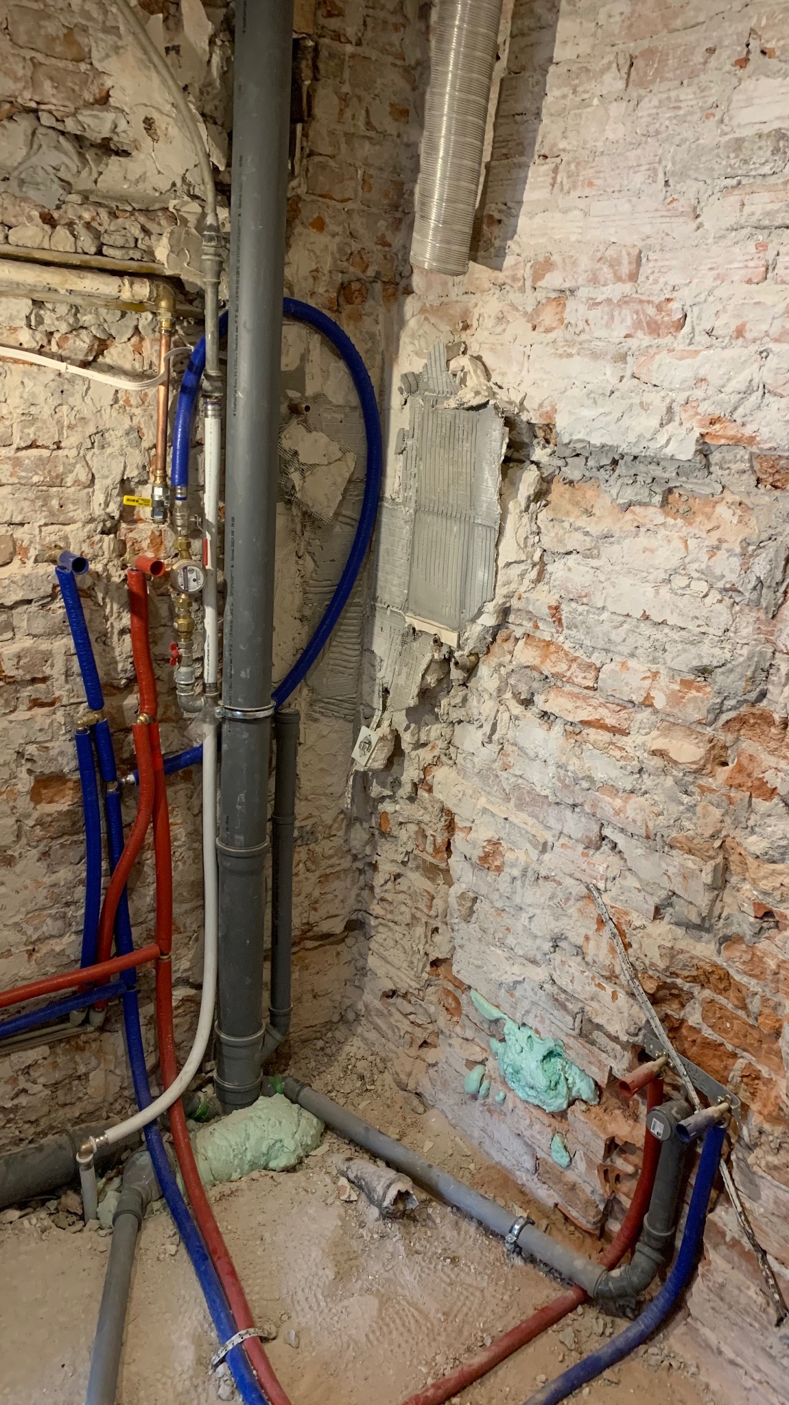 Fragment instalacji hydraulicznej w surowym wnętrzu z ceglaną ścianą. Widoczne rury miedziane, plastikowe (czerwone i niebieskie) oraz szare rury kanalizacyjne.