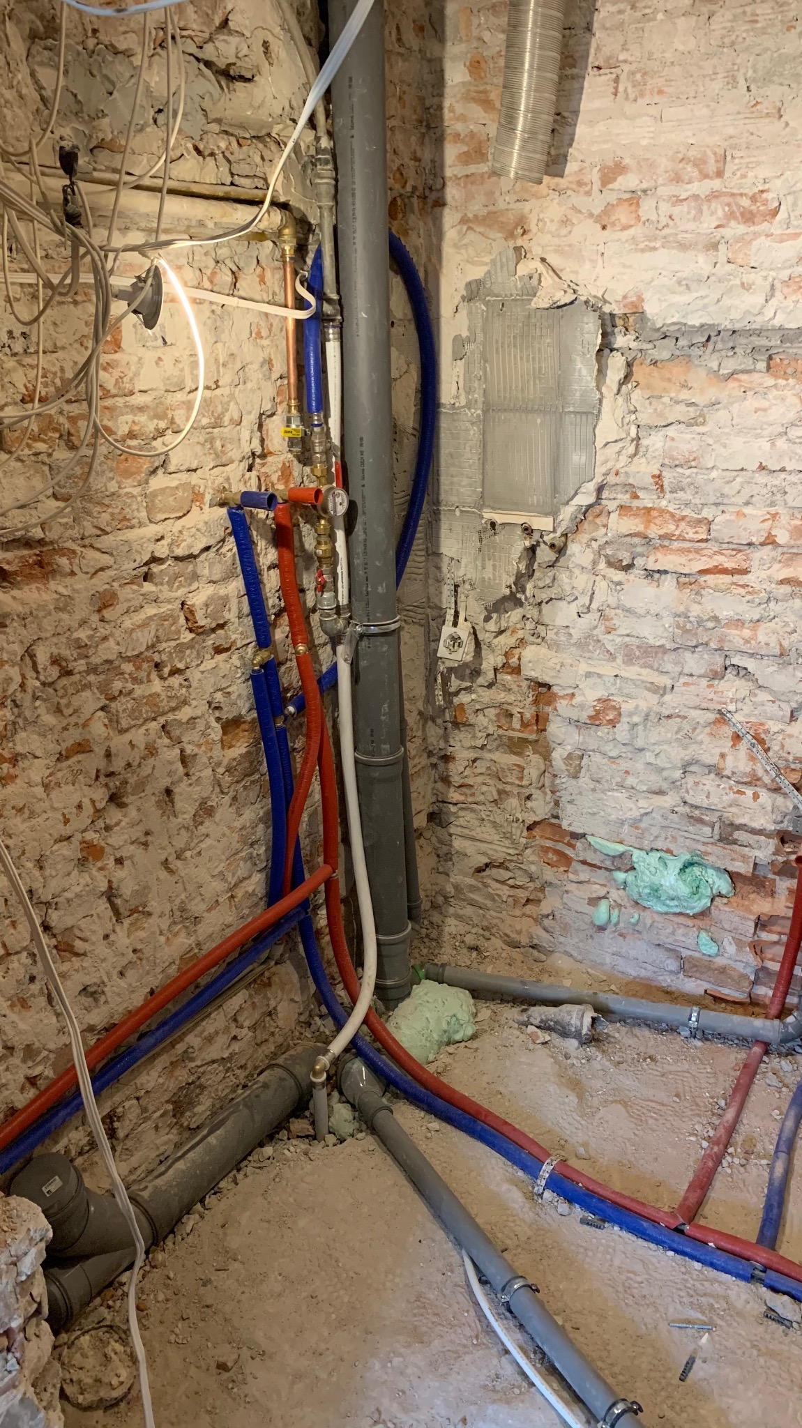 Fragment instalacji wodnej z rurami PEX (czerwone i niebieskie) oraz szarymi rurami kanalizacyjnymi na tle ceglanej ściany w trakcie remontu. Widoczne pianki montażowe.