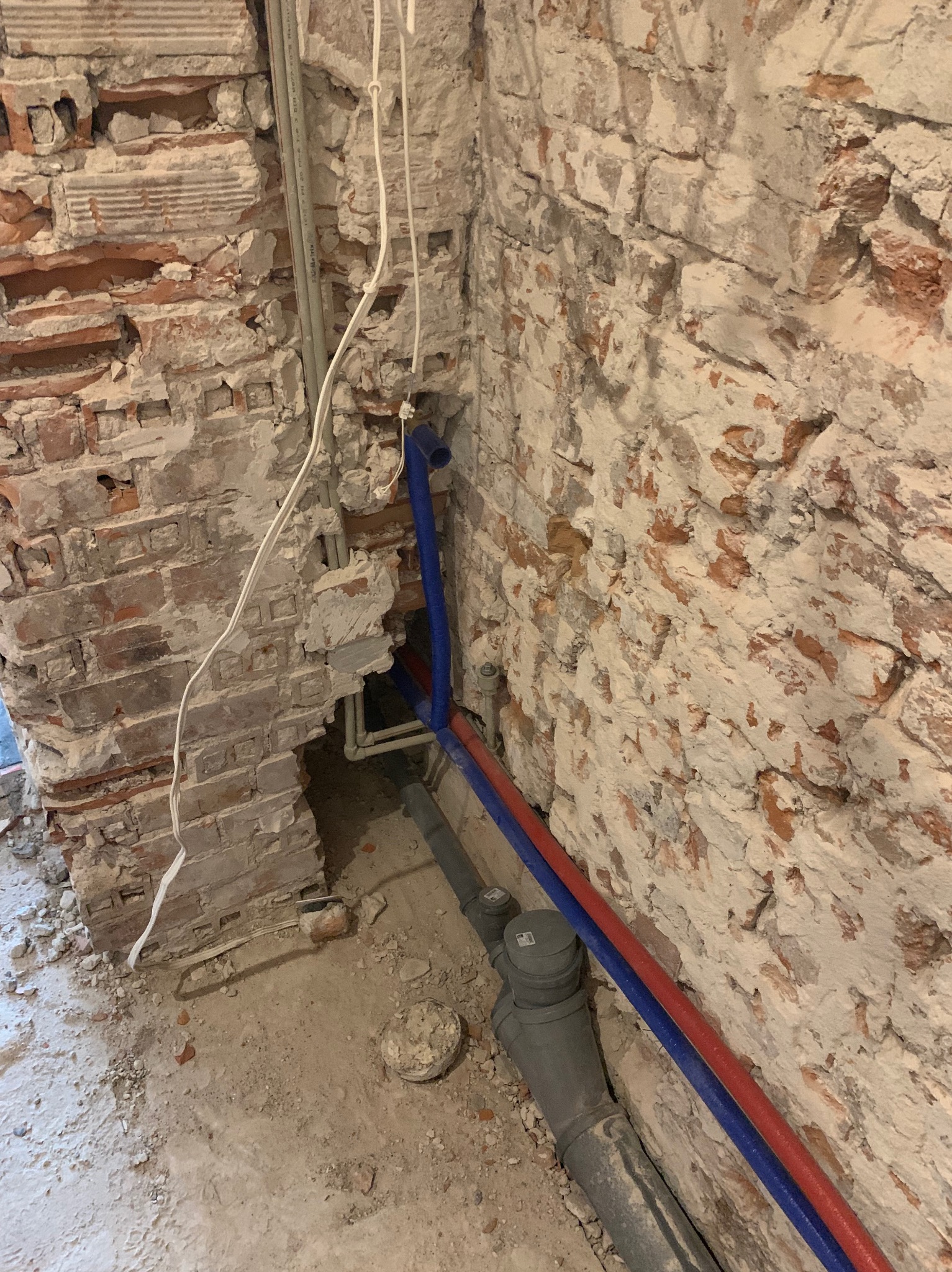 Fragment instalacji wodnej z rurami PEX (czerwona i niebieska) w surowym wnętrzu z ceglaną ścianą. Widoczne elementy kanalizacji i przewody elektryczne.