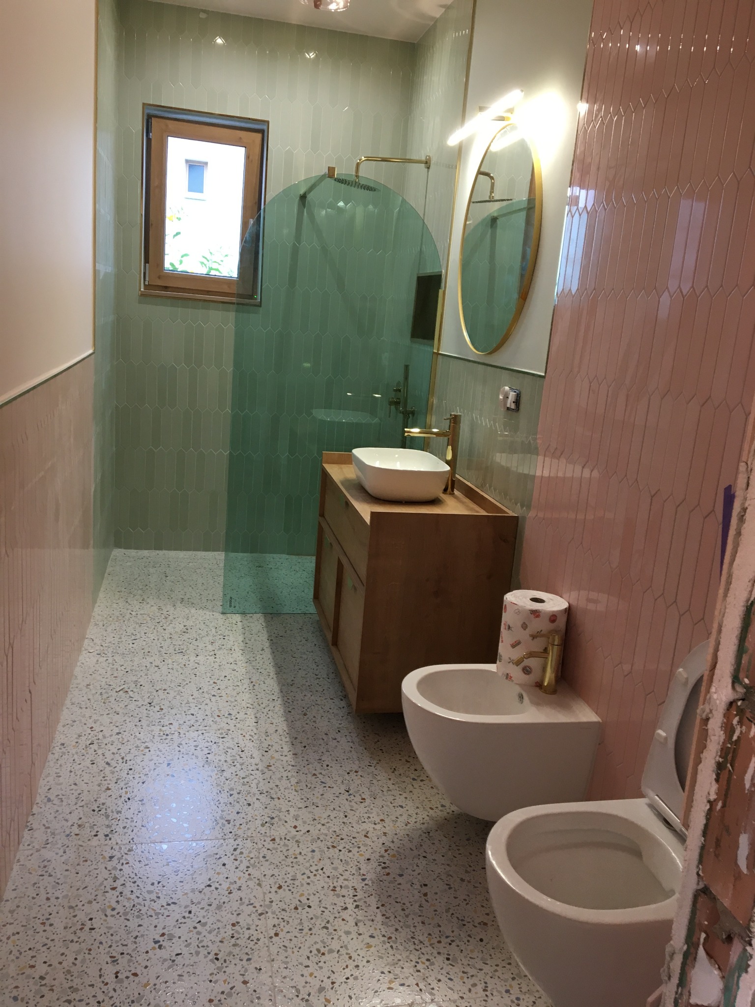 Wykończona łazienka z zielonymi i różowymi płytkami w kształcie łezki, drewnianą szafką pod umywalką, bidetem i toaletą, podłoga w stylu lastryko. Widoczne okno z drewnianą ramą i okrągłe lustro...