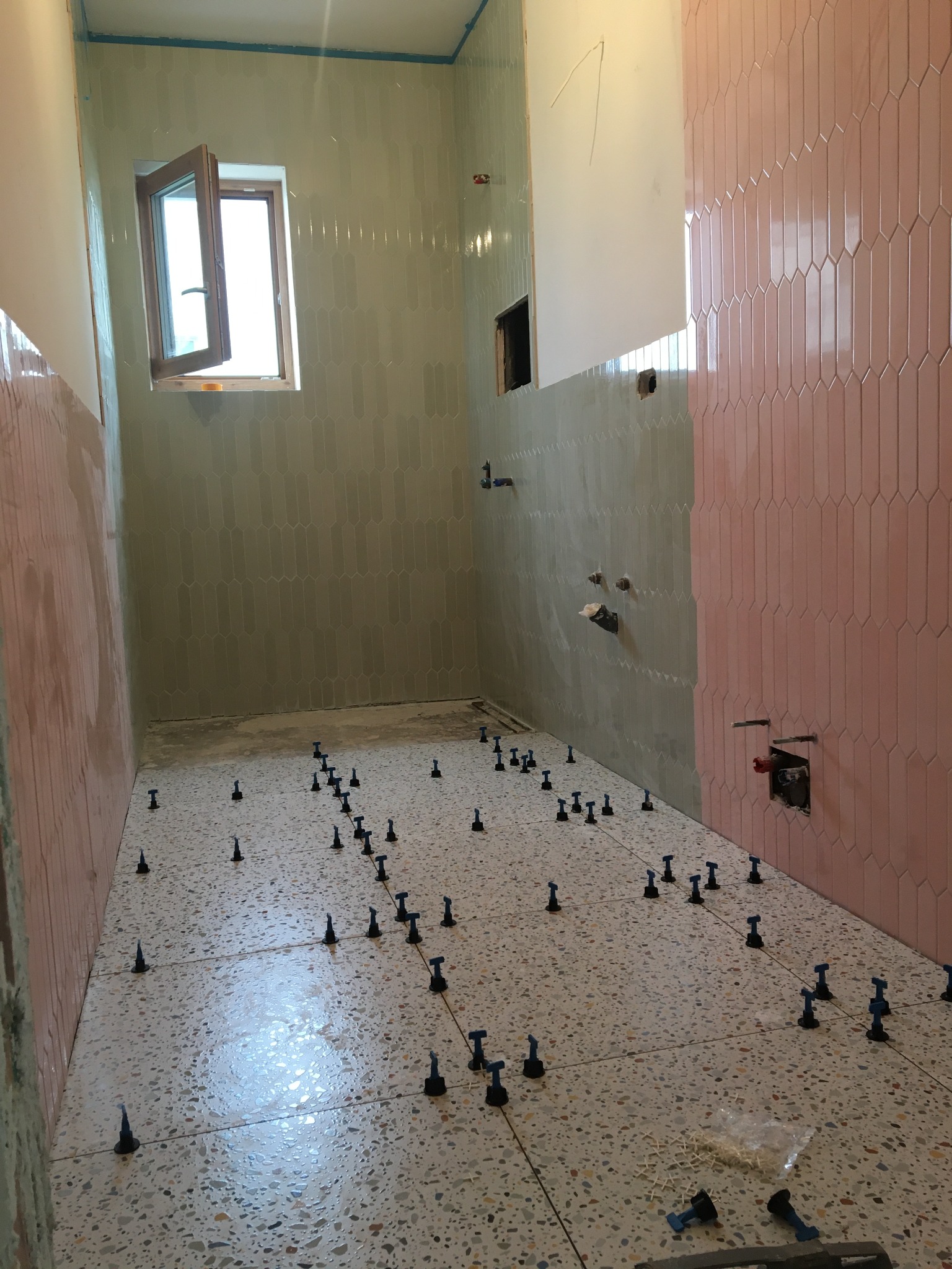 Wnętrze łazienki w trakcie prac wykończeniowych: ułożone płytki podłogowe z terazzo, pionowe płytki ścienne w odcieniach zieleni i różu, system poziomowania płytek, okno z otwartym skrzydłem.