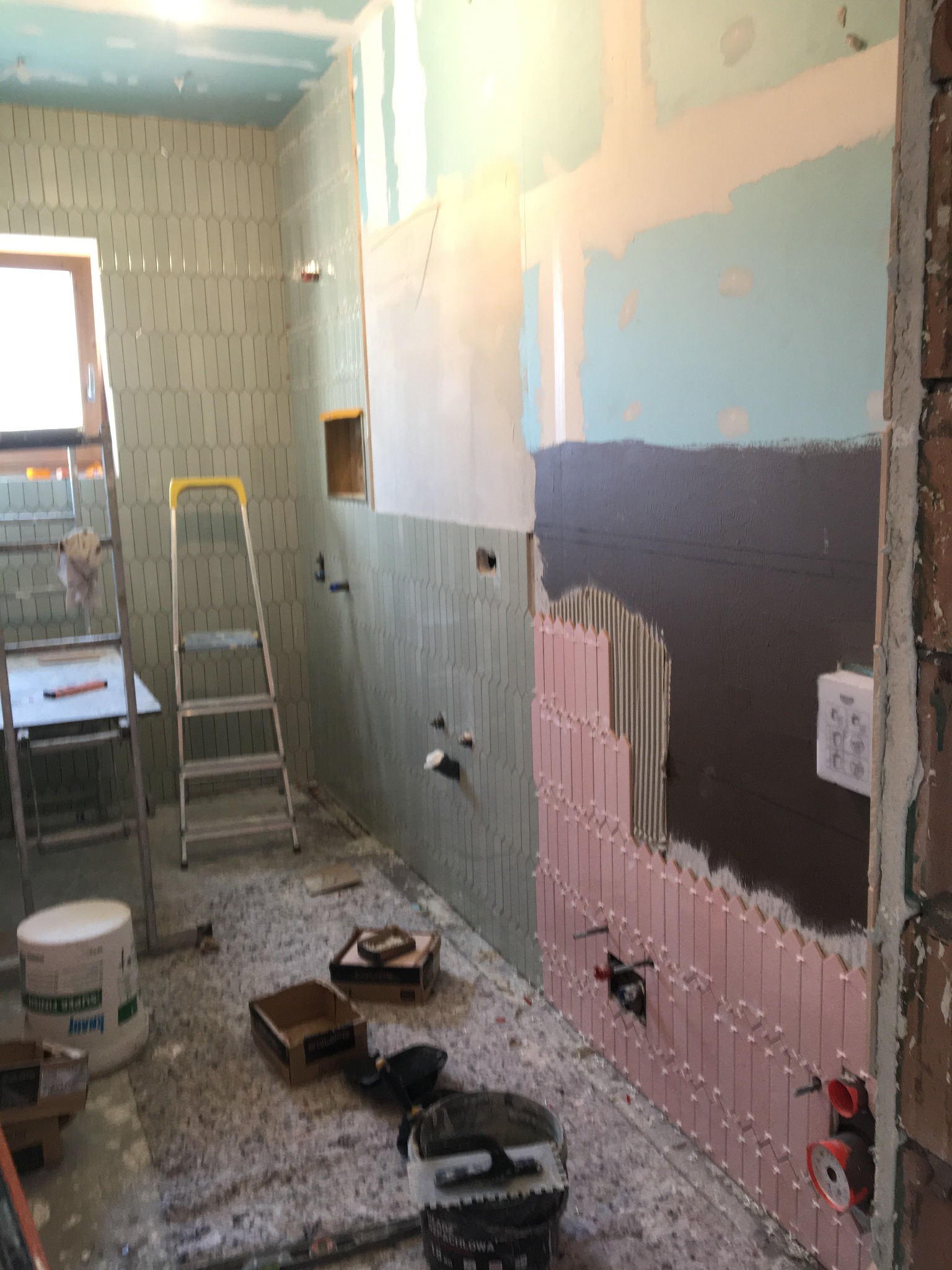 Wnętrze łazienki w trakcie prac: częściowo ułożone pionowe, podłużne płytki w odcieniach zieleni i różu, widoczne instalacje hydrauliczne, drabina i narzędzia na podłodze.