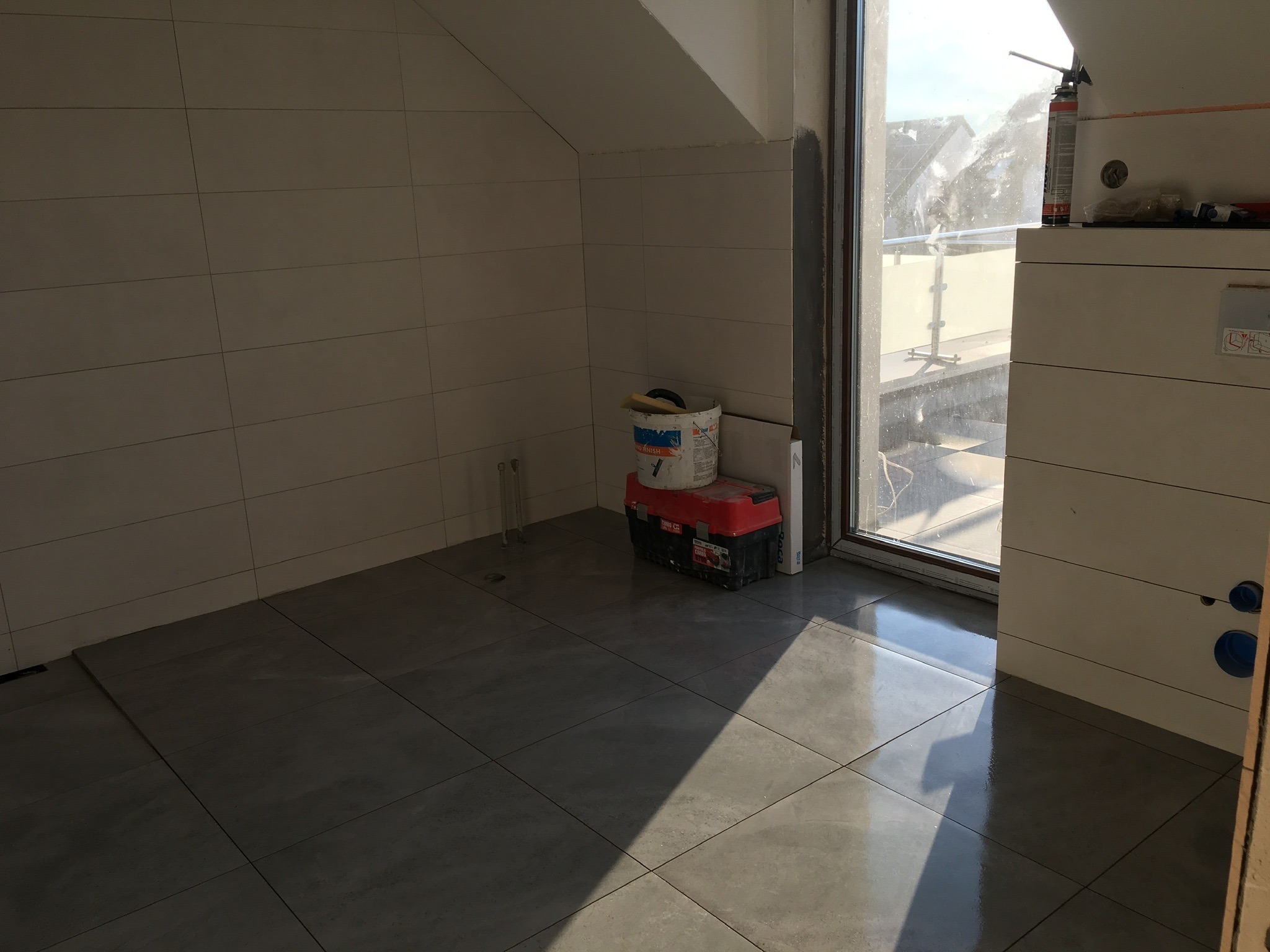 Wnętrze łazienki w trakcie remontu, z położonymi szarymi płytkami podłogowymi, częściowo wykończonymi ścianami z białych płytek, narzędziami i materiałami budowlanymi przy oknie.