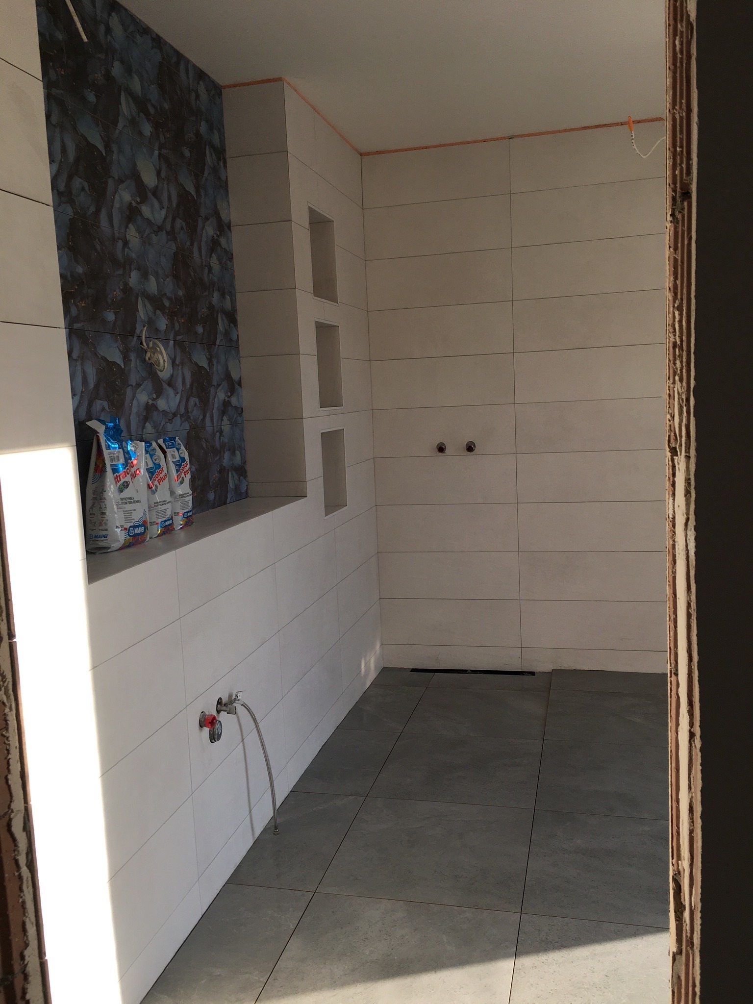 Wnętrze łazienki w trakcie wykańczania: ułożone szare płytki podłogowe i białe płytki ścienne z dekoracyjnym panelem imitującym marmur. Widoczne pakiety zaprawy klejowej Mapei, wnęki w ścianie...