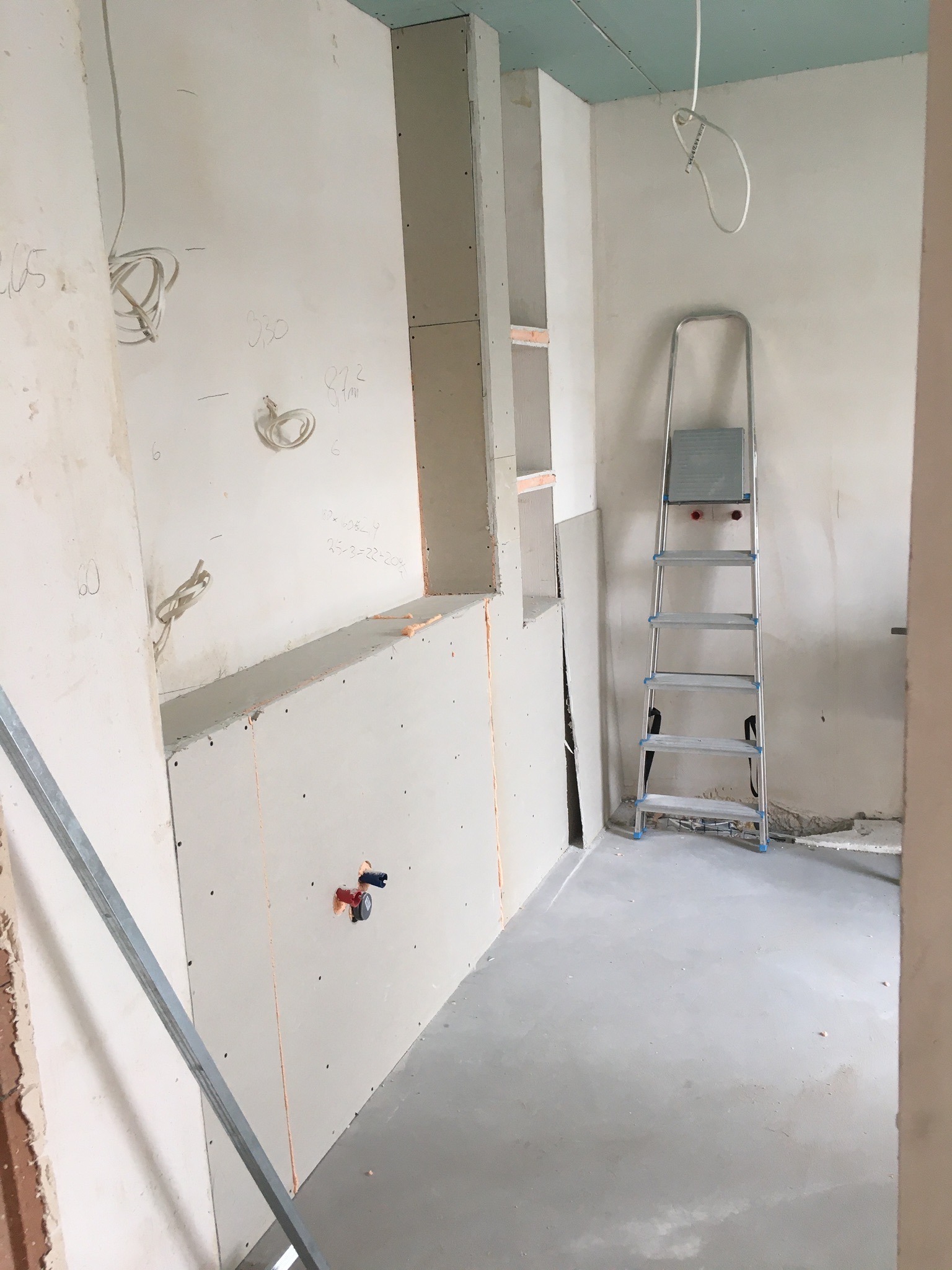 Wnętrze w trakcie prac: ściany wykończone płytami gipsowo-kartonowymi, widoczne instalacje, drabina aluminiowa, surowy beton na podłodze, notatki na ścianie.