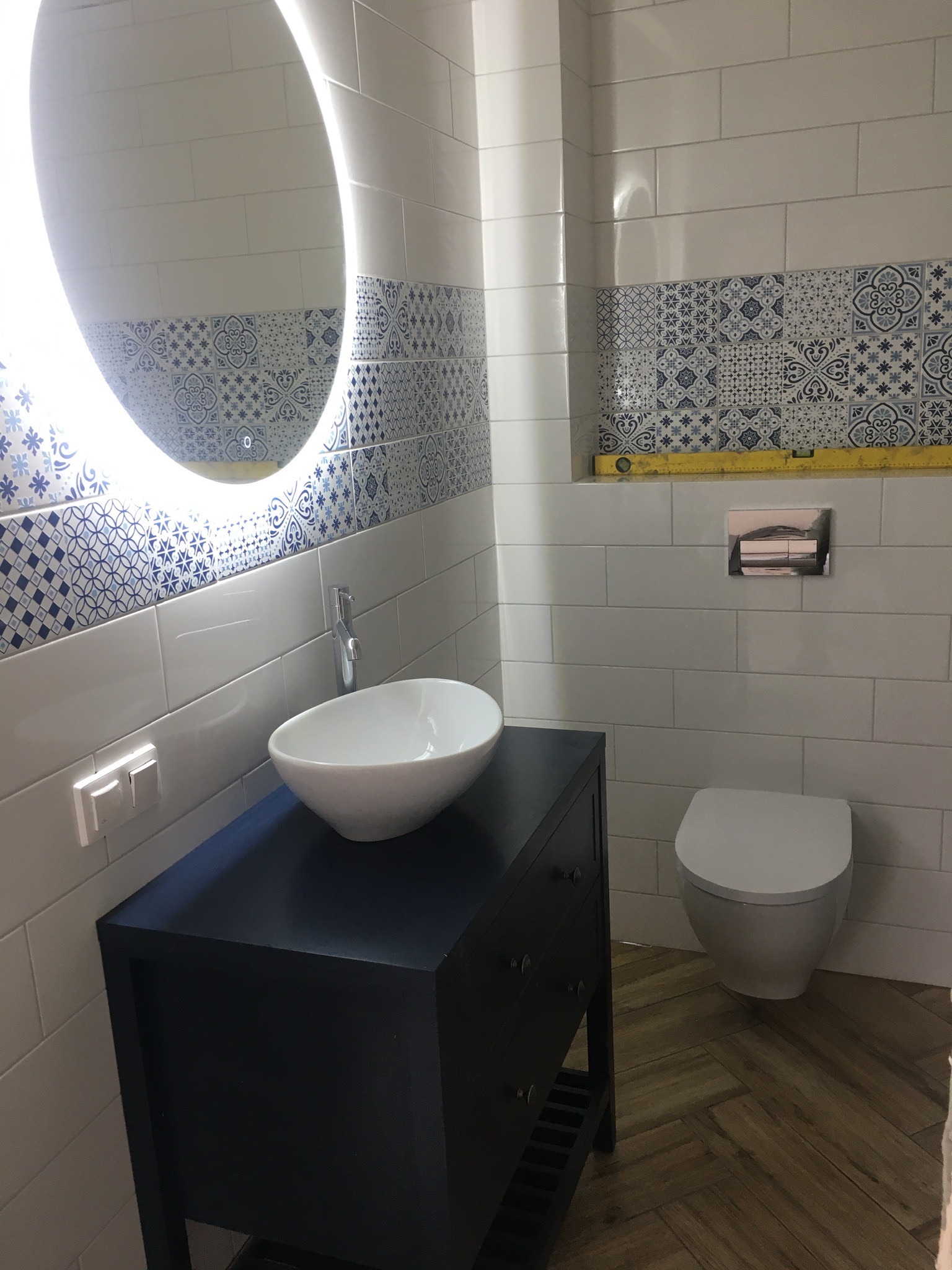 Wykończona łazienka z biało-niebieskimi płytkami dekoracyjnymi, owalnym lustrem z podświetleniem LED, umywalką nablatową na ciemnej szafce i toaletą w tle.