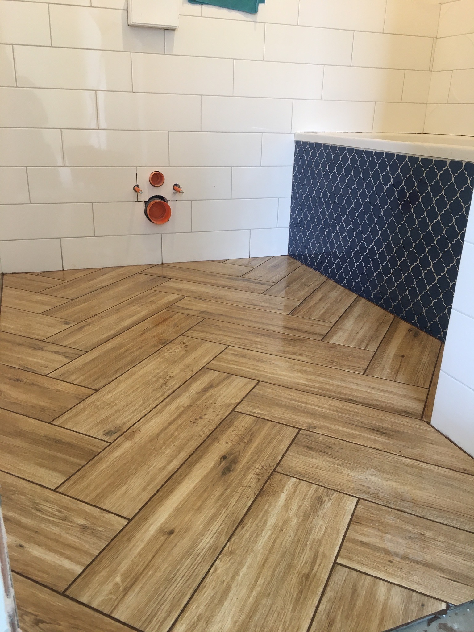 Ułożona podłoga z płytek imitujących drewno w jodełkę, białe kafelki na ścianie z widocznymi rurami i niebieska mozaika nad wanną w łazience.