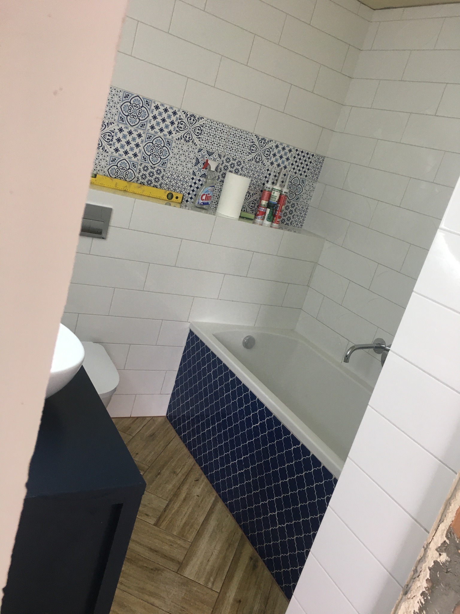 Wykończona łazienka z białą armaturą, granatową mozaiką na obudowie wanny i dekoracyjnym pasem płytek z niebieskim wzorem nad wanną, widoczna poziomica i środki czystości.