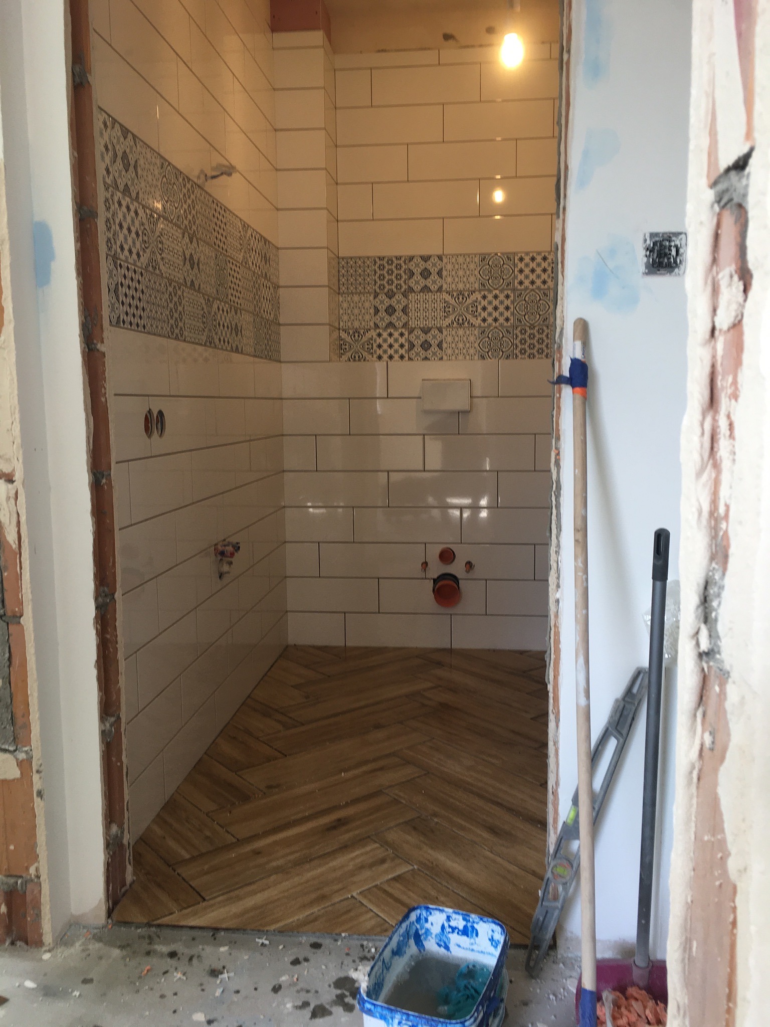 Wnętrze łazienki w trakcie układania płytek: białe płytki prostokątne na ścianach z dekoracyjnym pasem, drewnopodobna podłoga ułożona w jodełkę, widoczne narzędzia i wiadro z zaprawą.