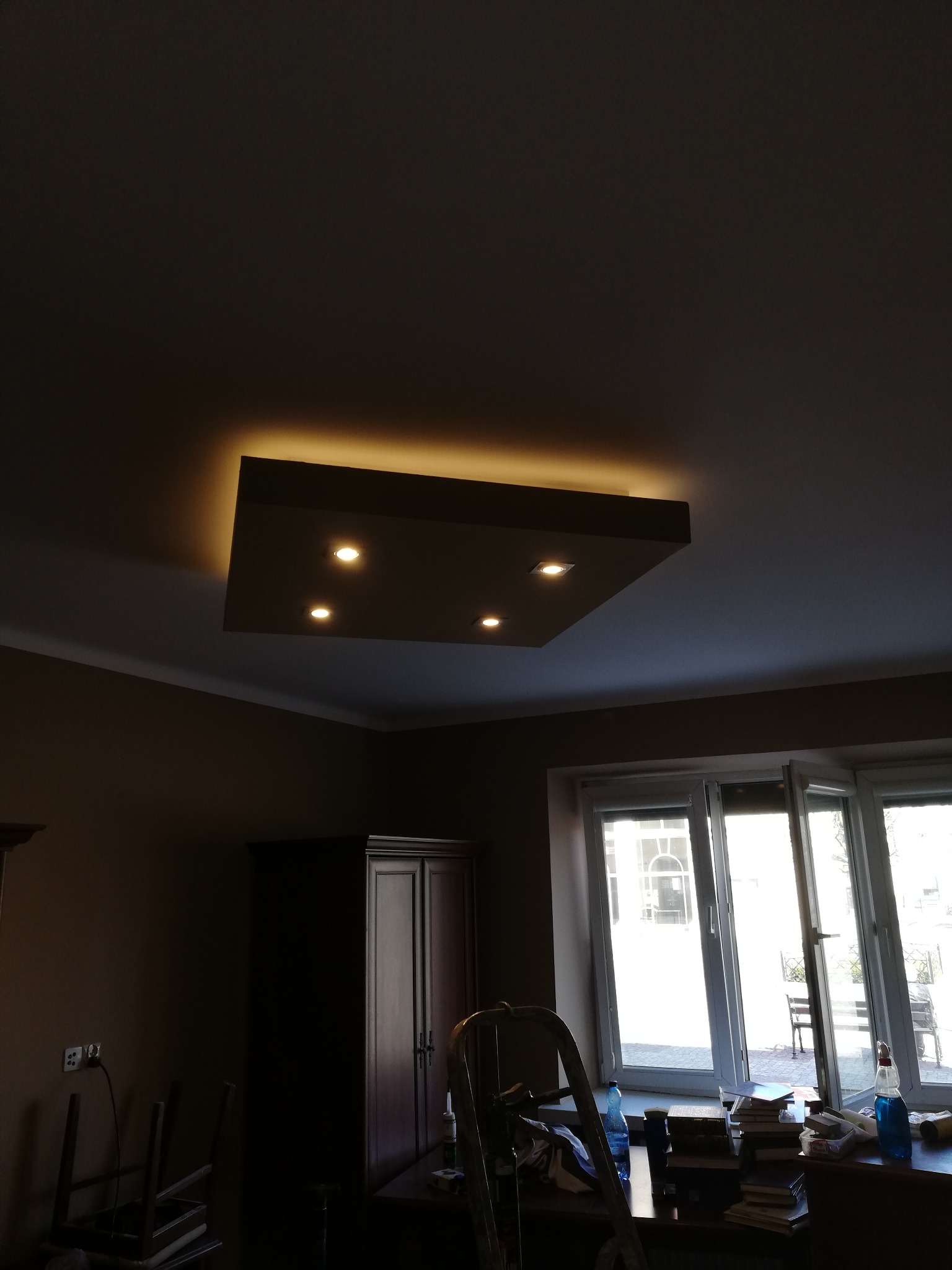 Sufit podwieszany z oświetleniem punktowym i taśmą LED w brązowym pokoju, widoczny fragment okna i mebli, drabina w tle.