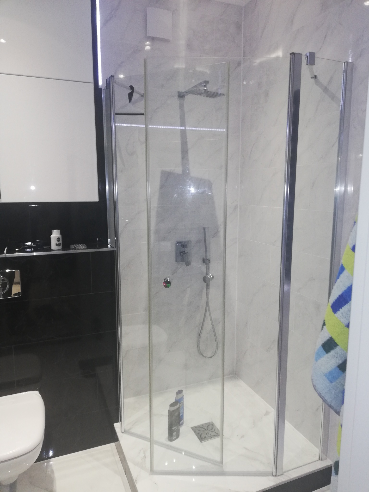 Nowoczesna kabina prysznicowa z deszczownicą, wykończona marmurowymi płytkami, widok na czarne płytki i białą toaletę.