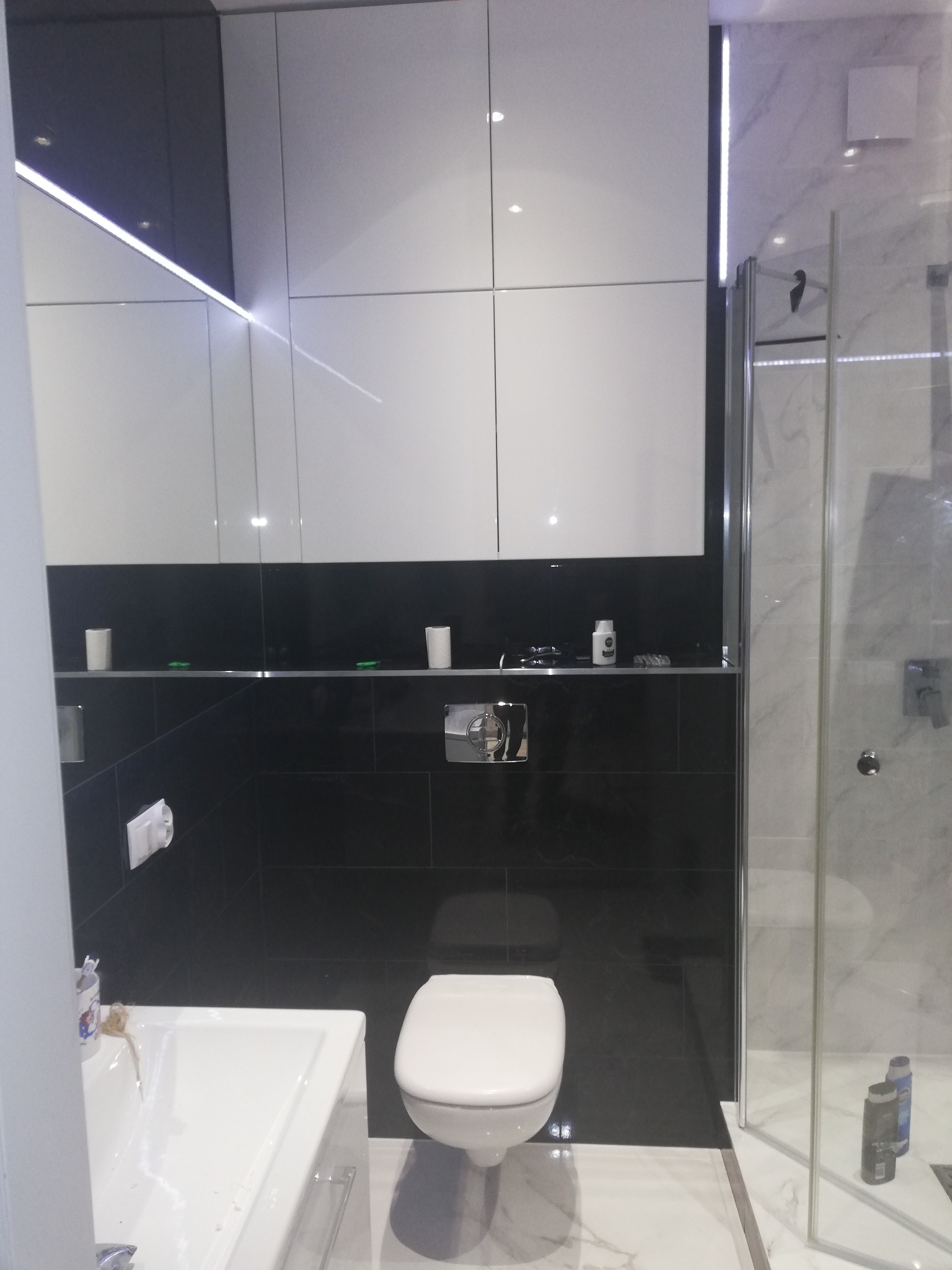 Nowoczesna łazienka z czarno-białymi płytkami, podświetlanymi szafkami, umywalką, toaletą i kabiną prysznicową z marmurowymi ścianami.