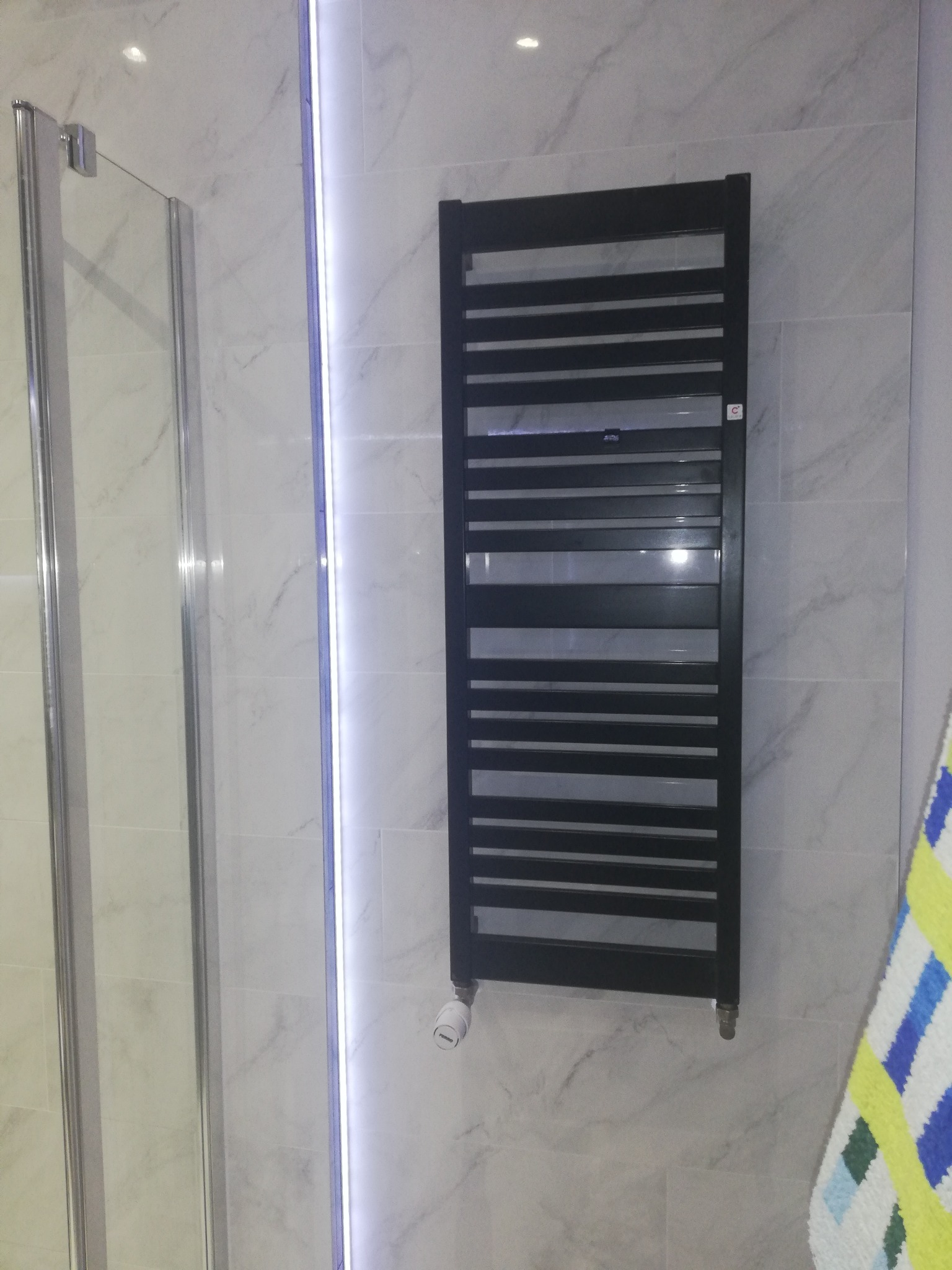 Czarny, drabinkowy grzejnik łazienkowy na marmurowej ścianie w Aleksandrowie Łódzkim, obok kabiny prysznicowej z oświetleniem LED.