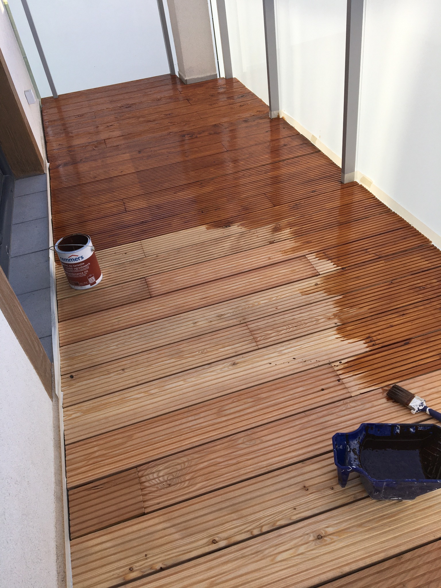 Olejowanie drewnianego tarasu: widoczna puszka z olejem do drewna, pędzel i kuweta malarska, część tarasu już zabezpieczona olejem, druga część jeszcze surowa.