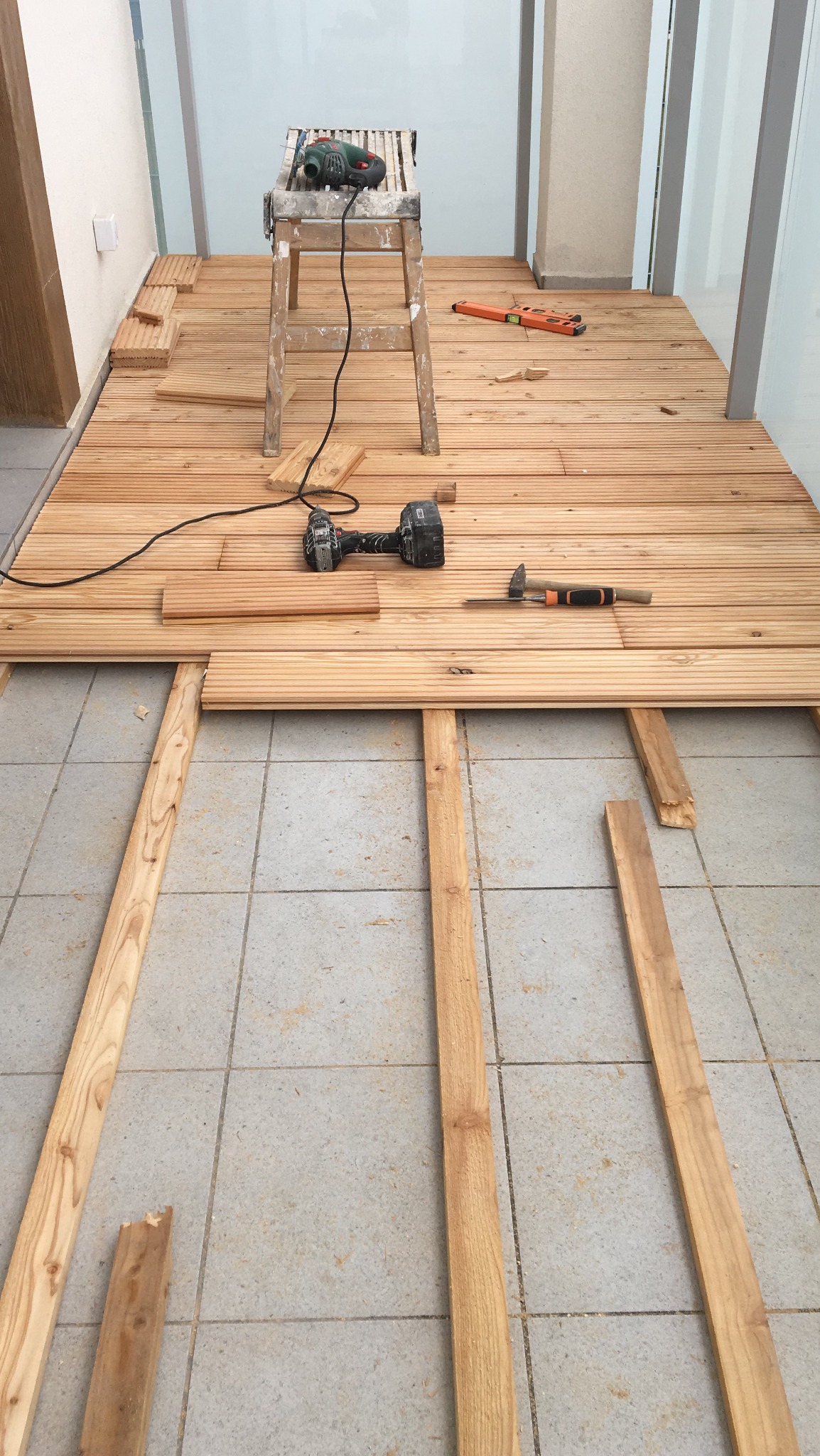 Montaż drewnianego tarasu na balkonie: deski ułożone częściowo na legarach, wiertarka i młotek na gotowej powierzchni, drabina z szlifierką w tle.