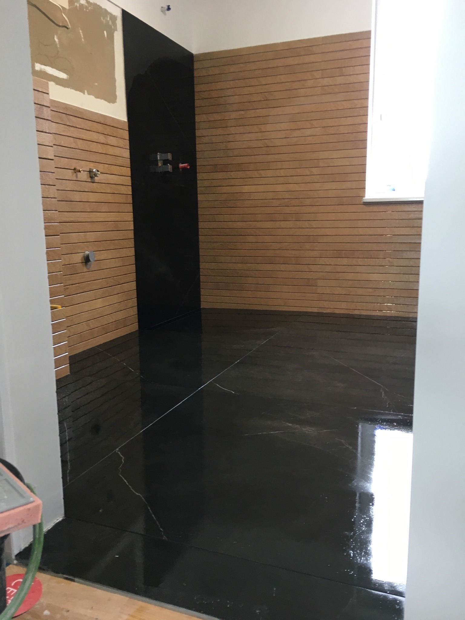 Wnętrze łazienki w trakcie remontu: drewnopodobne panele ścienne, czarna ściana z instalacją hydrauliczną oraz błyszcząca, czarna podłoga z efektem marmuru.