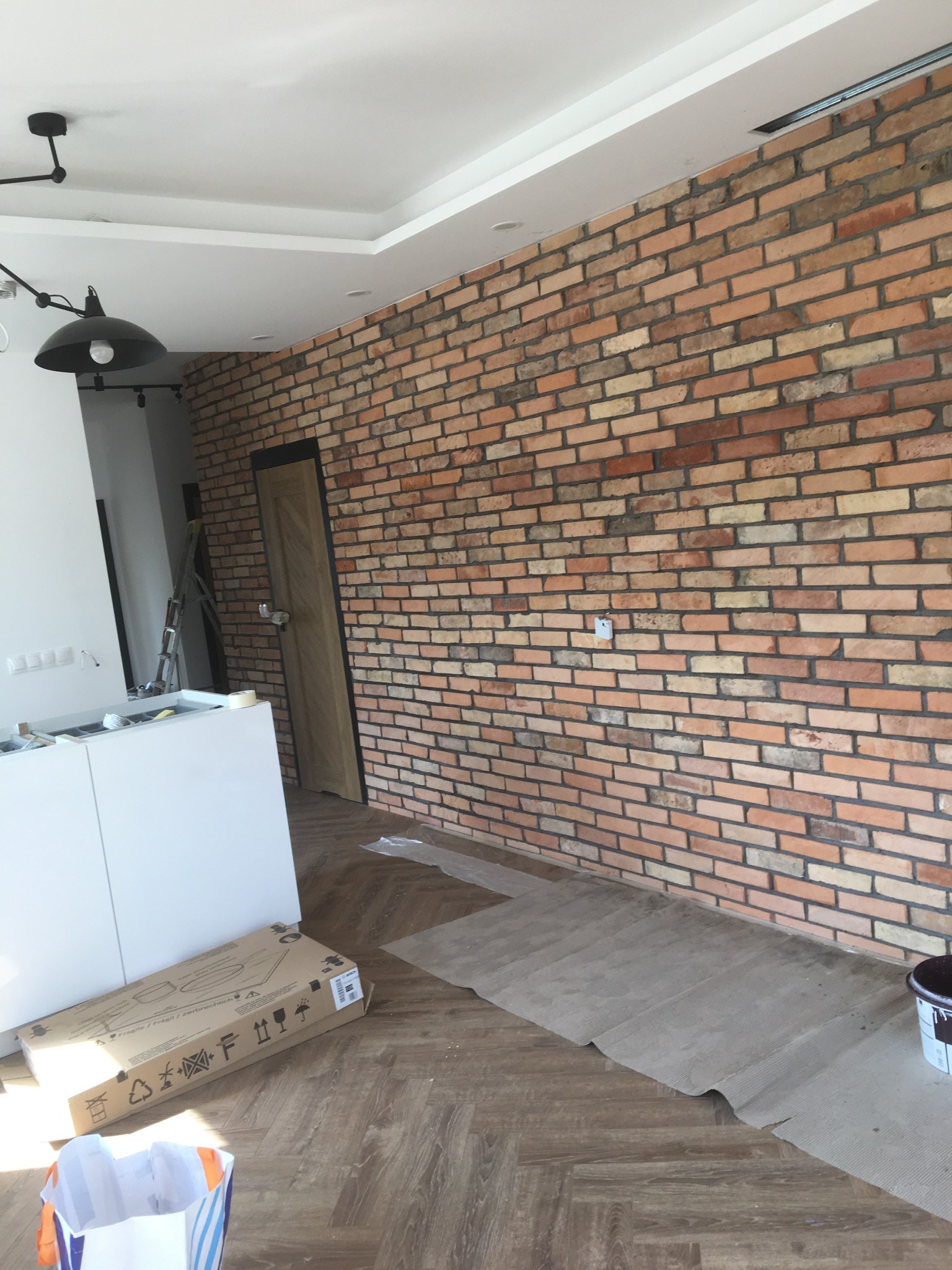 Ściana z cegły dekoracyjnej w jasnym wnętrzu, widoczna drabina, kartony i wiadro farby, podłoga w jodełkę.